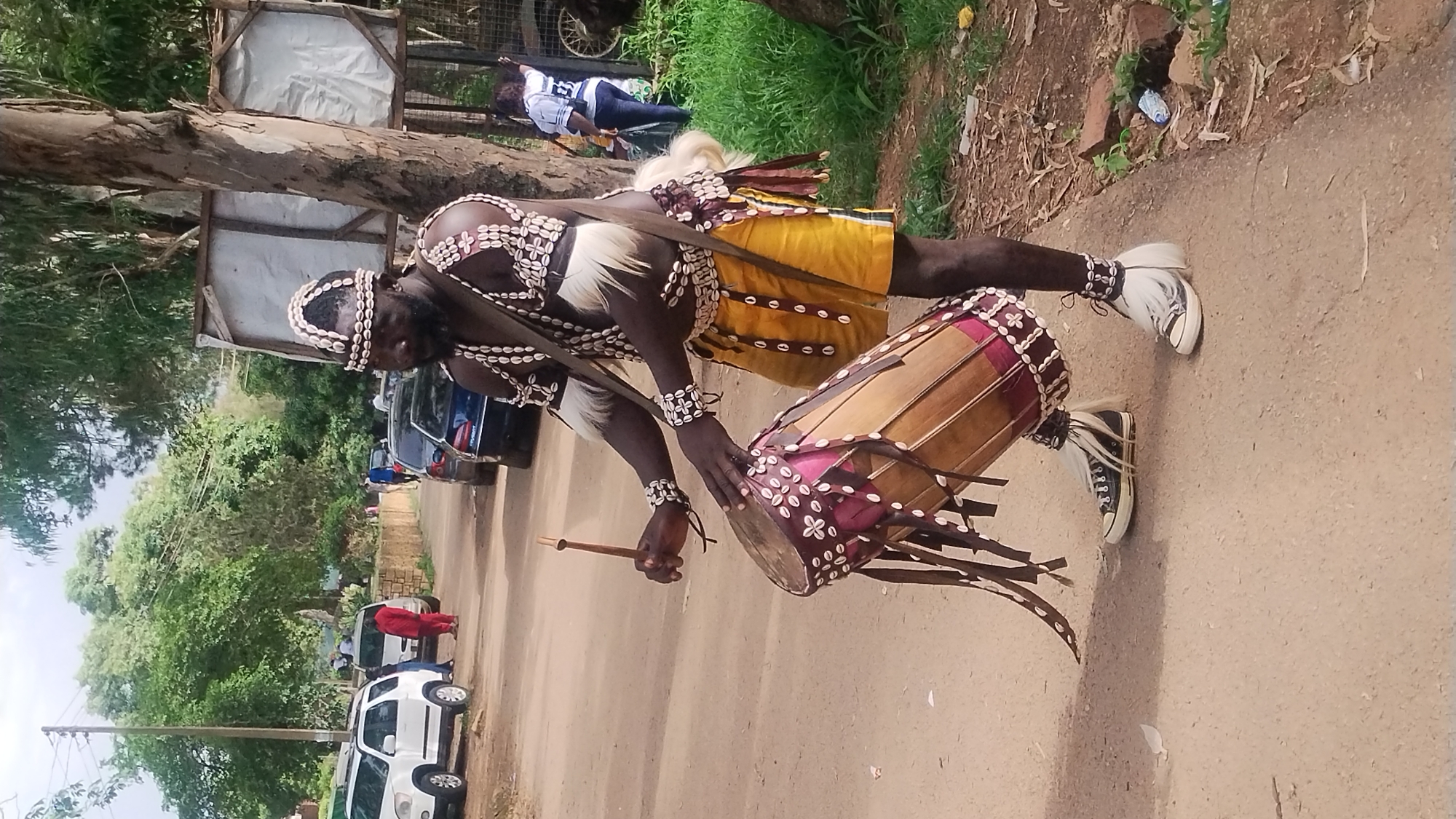A man in traditional attire plays a drum on a dirt road in Jos, Nigeria. The man, with dark skin and a beard, wears a beaded headdress, necklaces, armbands, and a belt adorned with cowrie shells. His outfit includes a yellow loincloth and white fur accents on his shoulders and ankles. He is in motion, holding a drumstick in his right hand and his left hand poised over a drum decorated with shells and fabric. The drum is cylindrical and rests on the ground. Black Converse sneakers with white fur adornments are visible on his feet. In the background, several cars, including white SUVs and dark sedans, are parked along the road. Trees line the left side of the road, and a simple wooden structure stands on the right, partially obscuring a person in a blue and white shirt. The ground is a mix of dirt and scattered debris.