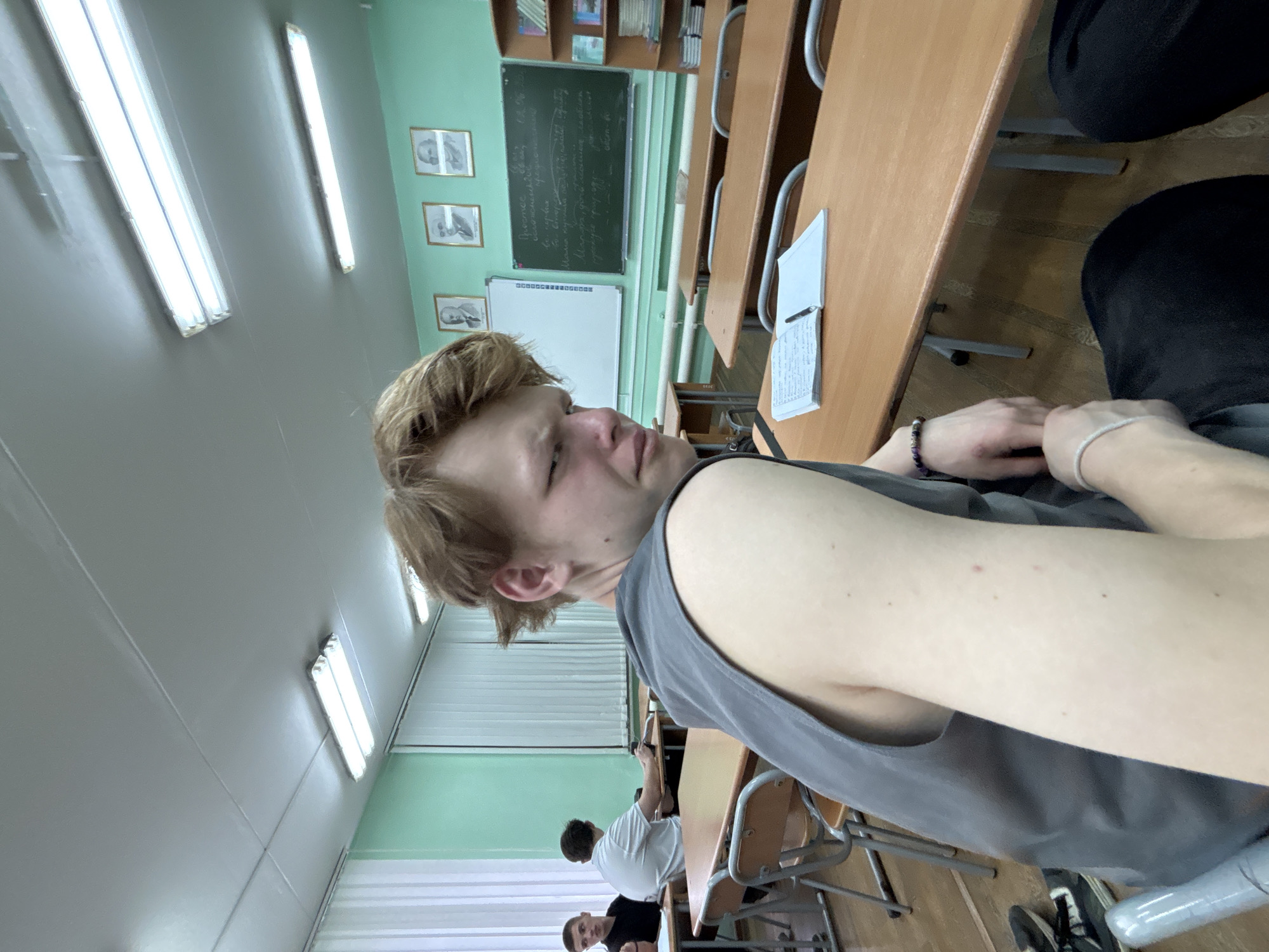 The image depicts a candid, slightly rotated view inside what appears to be a classroom or lecture hall. The main subject, a young male with short, light-colored hair, is positioned in the foreground, looking off to the left (viewer's right) with a relaxed expression, possibly a subtle smirk. He is wearing a sleeveless grey top and has bracelets on both wrists.
The setting is characterized by light green walls and a white ceiling equipped with multiple long fluorescent light fixtures. In the background, rows of wooden desks and chairs are visible, typical of an educational environment. Other students are present, seated at desks; one student wearing a white t-shirt and another in a black t-shirt can be partially seen.
Prominently displayed on the front wall are a dark chalkboard with white writing in what appears to be Cyrillic script, and a white whiteboard positioned below it. Three framed pictures, likely portraits or illustrations, hang on the green wall beside the chalkboard. On a desk in the mid-ground, an open notebook with written content and a pen are visible.
The scene conveys the atmosphere of a casual school or university class. The artificial lighting makes the time of day and external weather indistinguishable. No other significant text is clearly legible apart from the writing on the chalkboard.