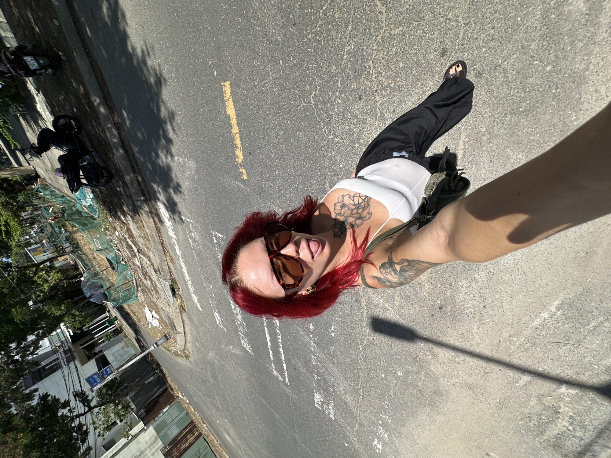 In Đà Nẵng, Vietnam, a young woman with bright red hair and large sunglasses is taking a selfie outdoors on a street. She is wearing a white tank top and black pants. She has tattoos on her arm and chest. The woman is standing near a crosswalk. In the background, there are buildings, trees, and two parked motorcycles. The scene appears to be a sunny day with shadows cast on the ground.