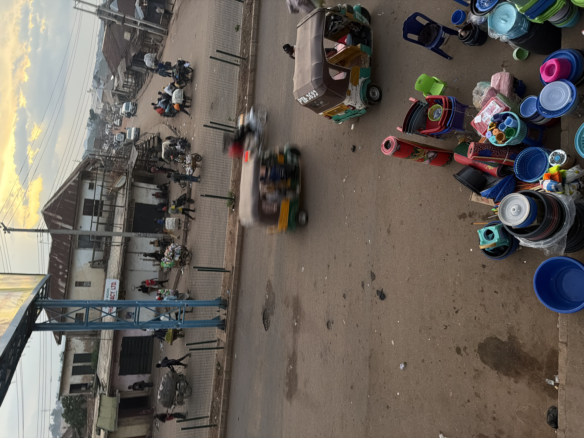 This media file depicts a busy street scene in Bukuru, Nigeria, likely during the late afternoon or early evening, given the warm, golden light of the setting sun illuminating the sky. The scene is characterized by a bustling market atmosphere with various modes of transportation and numerous objects scattered across the paved ground.
Prominently featured are several auto-rickshaws, commonly known as "Keke," used for transporting people and goods. One is parked with its license plate clearly visible as "PTA-3693." Another auto-rickshaw is in motion, its wheels blurred, suggesting the constant activity. Numerous individuals are visible, some walking on the sidewalk, others interacting near shops or with the vehicles. Their clothing and the general appearance of the environment suggest a typical day in a Nigerian urban or semi-urban setting.
The ground is cluttered with a variety of objects, predominantly plastic items such as buckets, bowls, and containers in various colors, including blue, green, and pink. Rolled-up mats, possibly for sale or storage, are also present, alongside what appear to be stacks of more household goods. Some of these items are arranged in piles, while others are scattered, indicating a marketplace or informal trading environment.
The setting includes buildings with corrugated iron roofs and a mix of weathered facades, suggesting a long-established area. A prominent blue structure, possibly a street light pole or a part of a larger canopy, stands on the left side of the frame, displaying a large advertising panel. A sign on one of the buildings reads "PHARMACY LTD."
The weather appears to be clear, with the sky filled with clouds catching the golden hues of the sunset, creating a warm and inviting ambiance despite the apparent busyness of the scene. The overall impression is one of a vibrant, active community engaged in commerce and daily life.