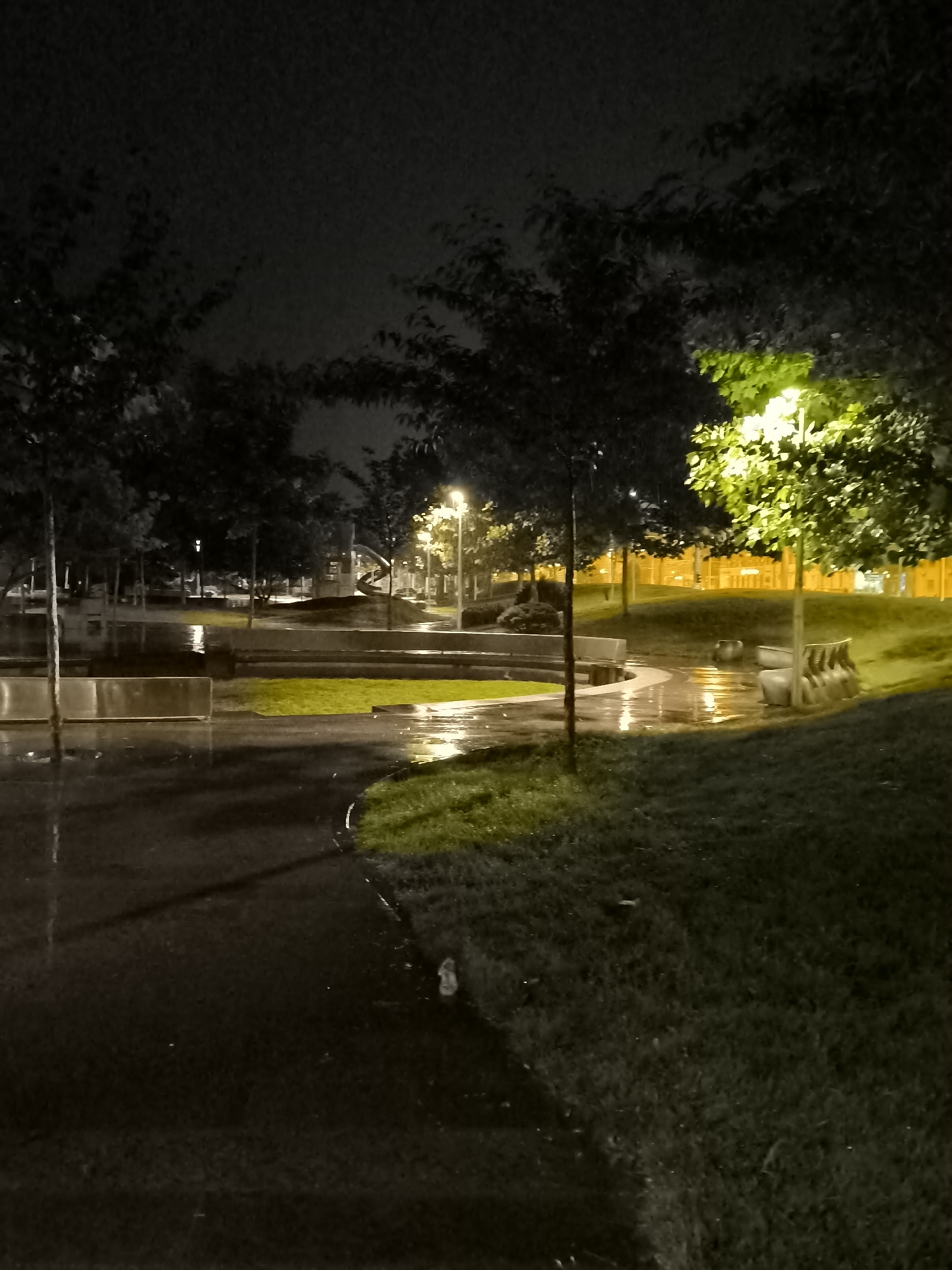 A nighttime scene in a park in Dnipro, Ukraine, with several trees silhouetted against a dark sky. A bright yellow lamppost illuminates a patch of green grass and the surrounding trees. The ground is wet and reflective, indicating recent rain. There are several concrete benches and planters visible, along with a paved pathway that curves through the park. In the background, a playground structure with a slide can be seen. The overall atmosphere is dark and somewhat eerie, with the artificial light creating stark contrasts.