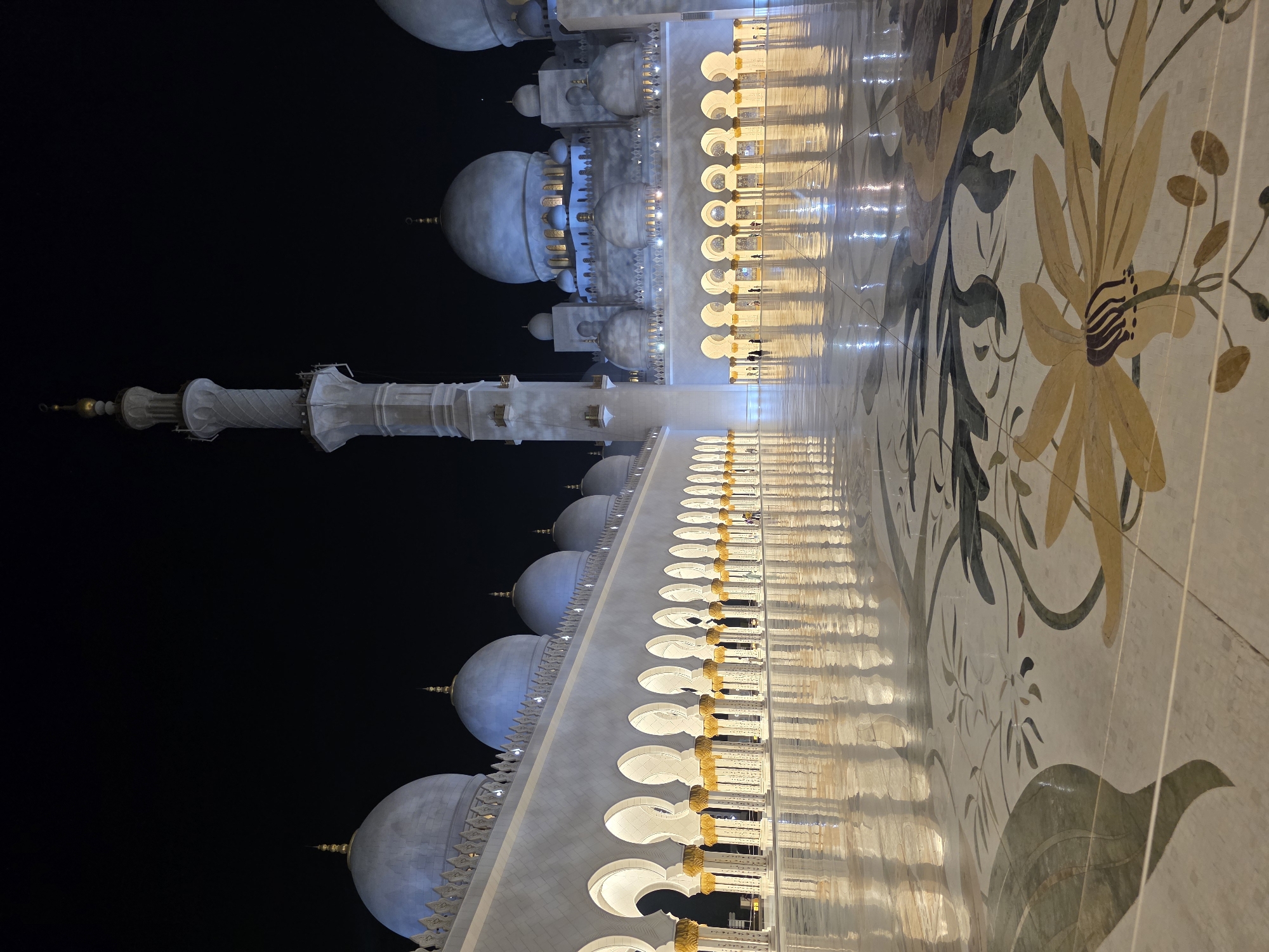 The image displays a night-time view of the Sheikh Zayed Grand Mosque in Abu Dhabi, United Arab Emirates. The mosque's exterior, characterized by its white marble domes and minarets, is illuminated, casting reflections on the polished floor. The foreground features intricate floral inlays on the floor, composed of marble and other stones in shades of green, yellow, and brown. In the middle ground, a colonnade of arches with ornate golden columns leads the eye towards the main structure of the mosque. The main building exhibits multiple domes of varying sizes, with the largest central dome prominent. A tall, slender minaret rises vertically in the mid-left of the frame, its textured surface clearly visible. The sky is dark, indicating it is nighttime. A few individuals, appearing as small figures in silhouette, are present in the middle ground, walking or standing within the mosque's courtyard. The overall scene conveys a sense of grandeur and architectural splendor.