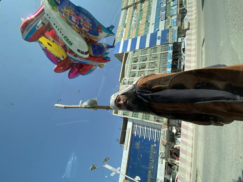 The image depicts a street scene in a commercial district of Kandahar, Afghanistan, observed through a reflective surface. An adult male, approximately center-frame, with a dark beard, wears a light-colored head wrap and a brown, patterned outer garment over a dark vest. He looks directly forward. To his upper right, a dense cluster of colorful foil balloons, many shaped like airplanes and other vehicles (one displaying "SUPER TEAM"), ascends into the clear blue sky.
In the background, to the left, stands a modern multi-story building with a blue glass facade, prominently labeled "COMMERCIAL CENTER" in English, with additional text likely in local script. Further right, a row of older, multi-story buildings lines the street, featuring various architectural styles, including one with distinctive blue and white vertical stripes.
A paved road occupies the lower portion of the frame, separated from pedestrian areas by a low concrete barrier painted with red and white diagonal stripes. Partial views of vehicles are discernible on the road and parked alongside. Streetlights with multiple LED panels are mounted on tall poles on the left. A rudimentary wooden pole with a plastic bag tied to it is visible behind the central figure. The sky is clear and blue.
