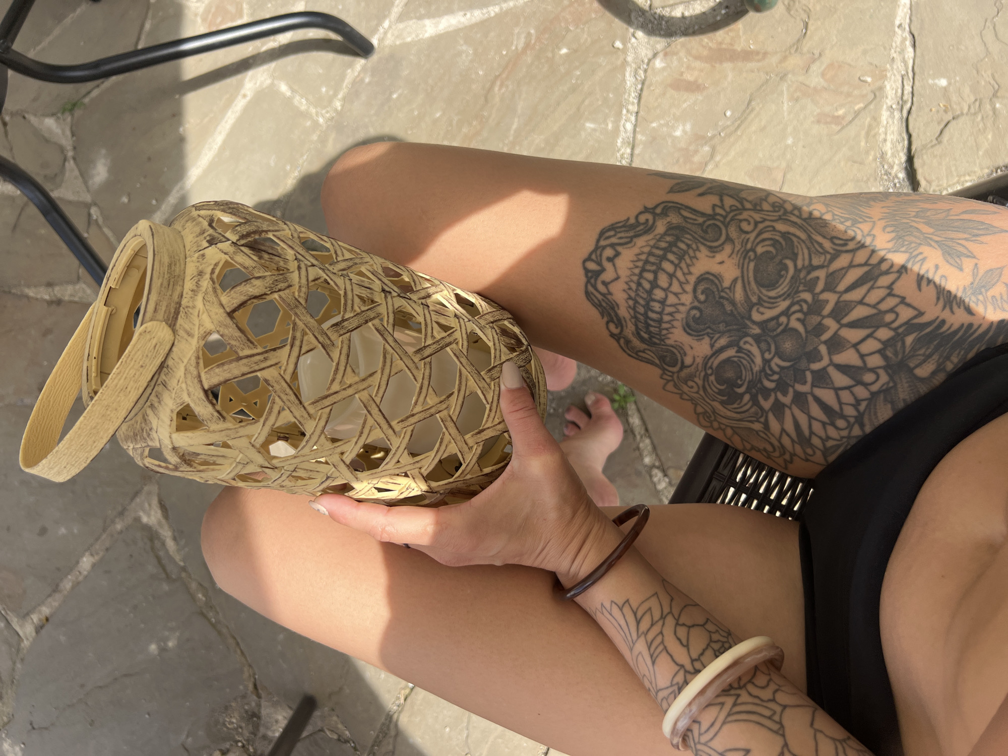 A tanned person with tattoos on their legs and arms sits on a chair outside on a sunny day. The person holds a decorative, woven candle holder in their left hand. A glimpse of the person's foot is visible. The background features stone paving and a chair leg. The location is Boguchar, Russia.
