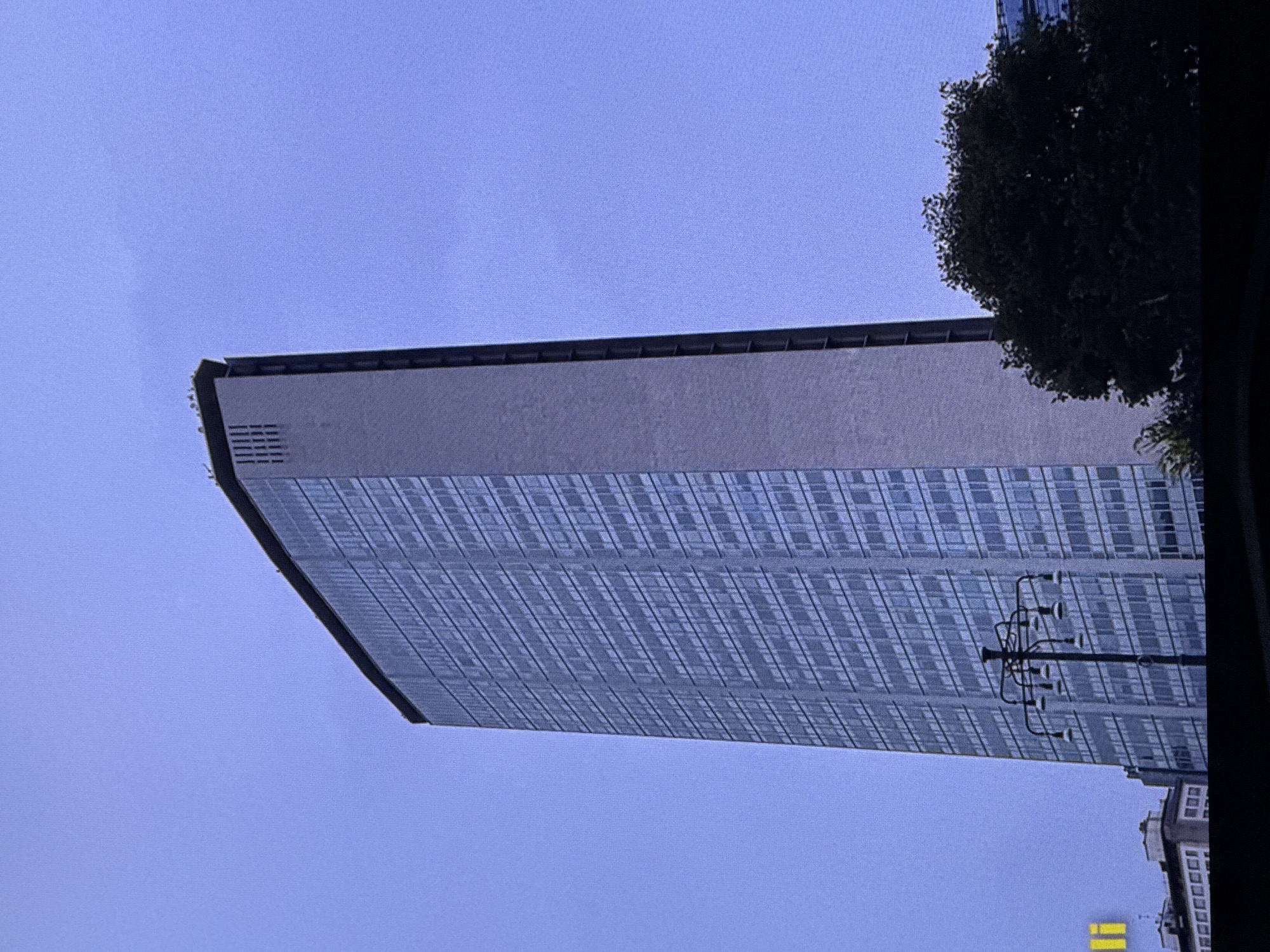This is a full-frame, low-angle shot of the Pirelli Tower, a skyscraper located in Milan, Italy, not Dubai. The building is made of concrete and glass and features a distinctive tapering shape. The sky is a pale blue, and there are no people or other significant activity visible in the frame. A streetlamp with multiple lights stands in front of the tower, and a dark, leafy tree is visible to the right. The lower portion of the image is obscured by a black bar.