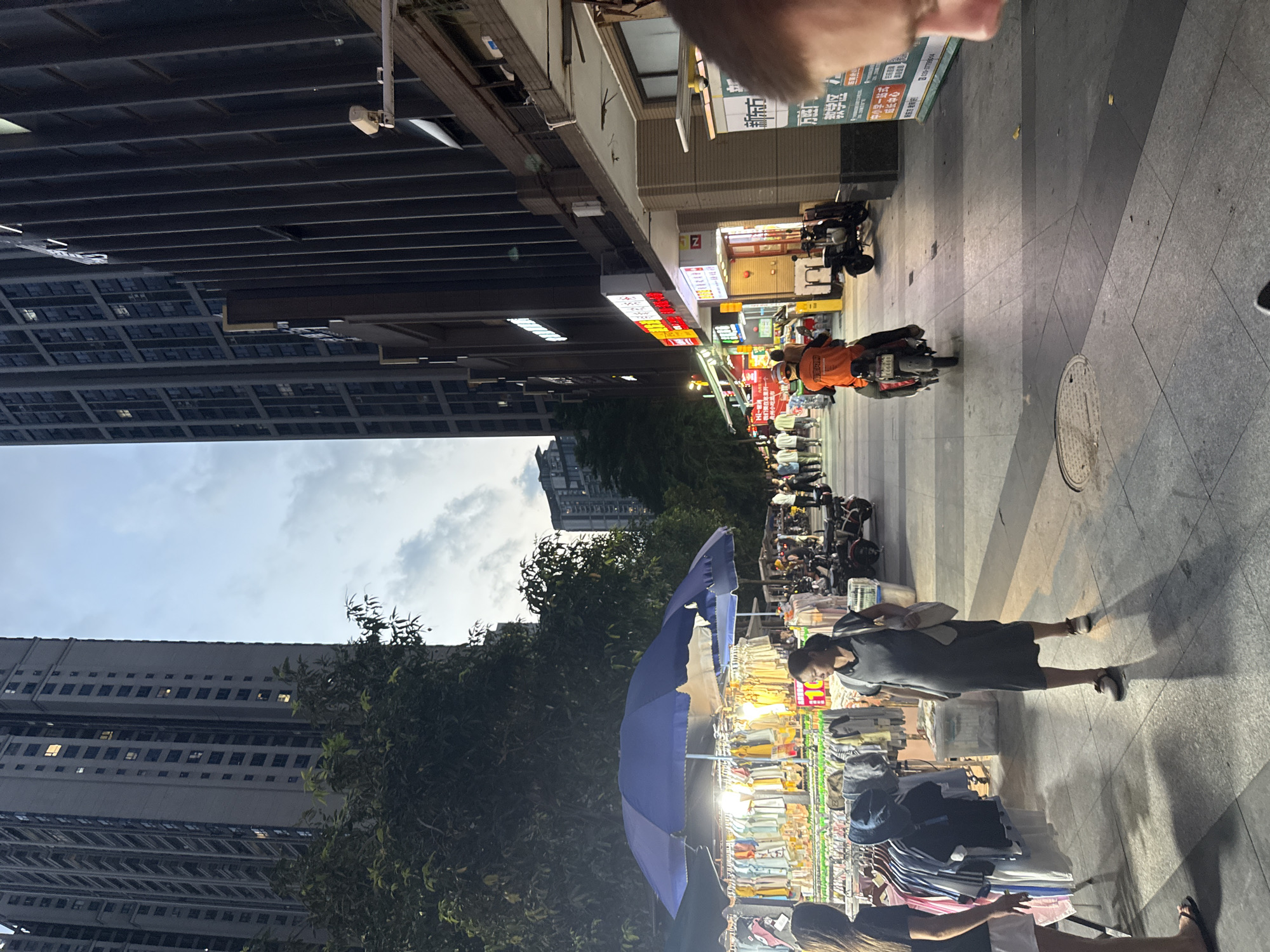 This media file captures a street scene in Haizhu District, China, likely during the late afternoon or early evening. The orientation of the image suggests a person is holding the camera and looking down at the scene. The setting is a bustling marketplace along a pedestrian walkway, with a large building on the left and shops lining the right side of the street.
Several people are visible, including a person on a scooter wearing an orange jacket, and shoppers browsing various stalls. One woman in a dark dress is prominently featured, looking towards the market. Another person, partially visible, is seen from behind, also near the market stalls. The stalls themselves are filled with clothing and other merchandise, displayed on racks and tables, with a large blue umbrella providing shade over some of the goods. Various motorcycles and scooters are parked along the walkway, indicating a common mode of transportation in the area.
The time of day appears to be transitioning from daylight to dusk, as indicated by the lighting and the sky, which shows a mix of clouds and a soft sky. The environment seems urban and active, with the architecture of the buildings suggesting a commercial or residential area. The presence of street vendors and a high volume of foot traffic points to a lively commercial district. There is some visible text on signs and banners, but it is mostly illegible in this resolution. The overall mood is one of everyday commerce and urban activity.
