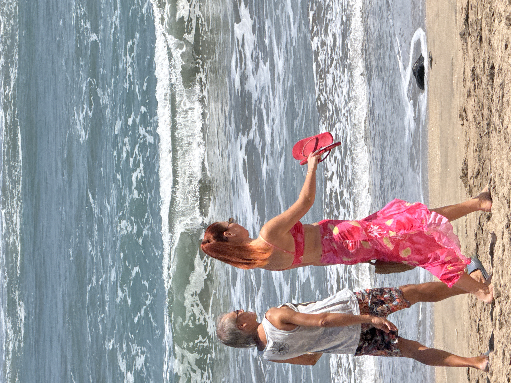 In Kuta, Indonesia, two individuals, a man and a woman, are walking on a sandy beach. The man, wearing a white tank top and patterned shorts, has grey hair and is looking to his left. The woman, with reddish-orange hair styled in a ponytail, is wearing a pink bikini top and a vibrant pink sarong with yellow accents. She is holding a red flip-flop in her right hand and appears to be looking towards the water. A woven bag is slung over her left shoulder. The ocean behind them features moderate waves with white foam. A dark, partially submerged rock is visible in the shallow water near the shore. The scene is captured on a bright, sunny day.