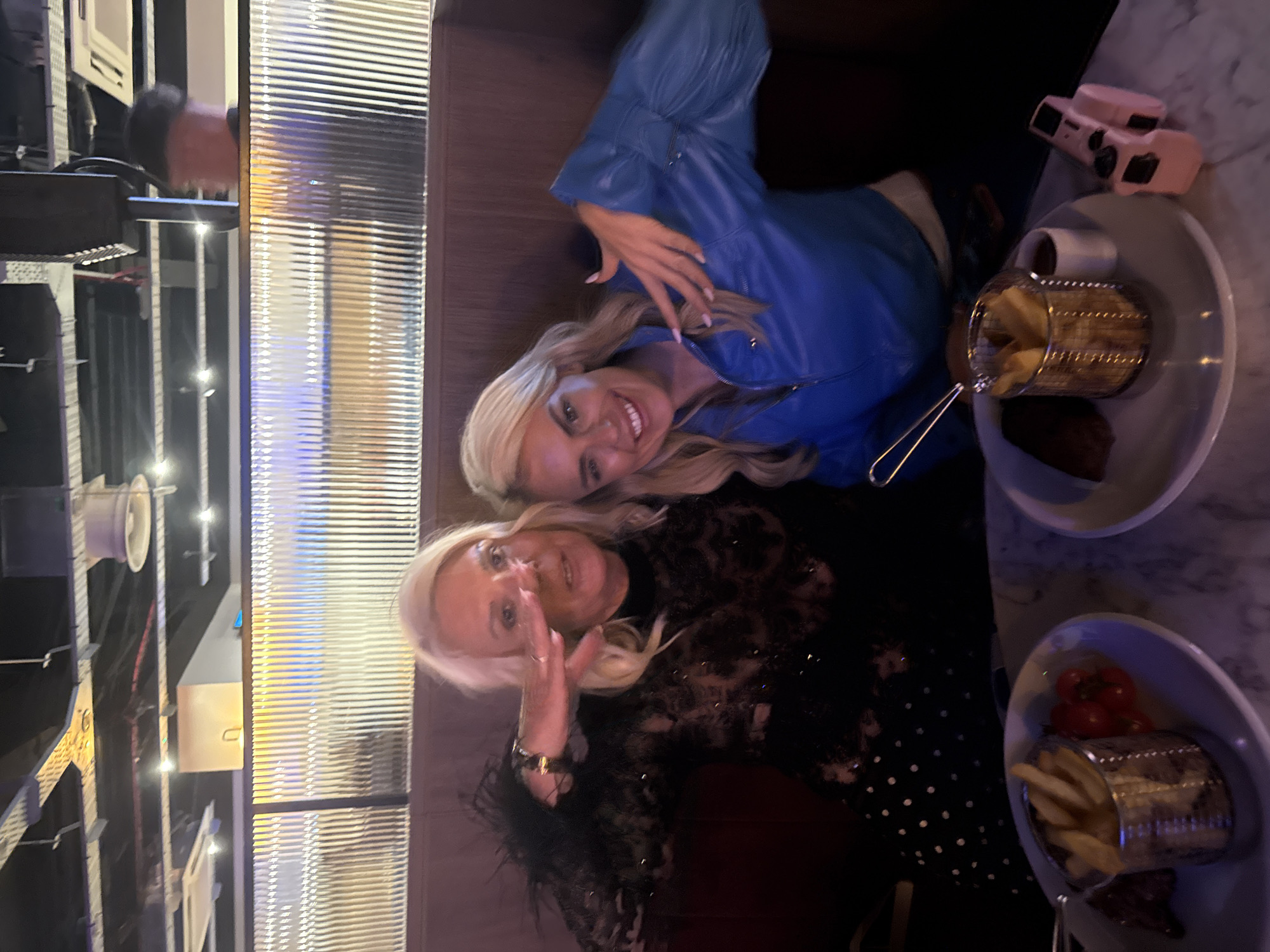 Two women are seated at a table in a dimly lit restaurant, with a patterned partition behind them and exposed ceiling infrastructure above. The woman on the left, with blonde hair, is wearing a black, sheer, sequined dress with feathered sleeves and is holding her hand up to her face. The woman on the right, also blonde, is wearing a blue jacket and smiling. In front of them are plates with food, including fries in metal baskets, and small cups of sauce. A pink object, resembling a camera, is on the table.