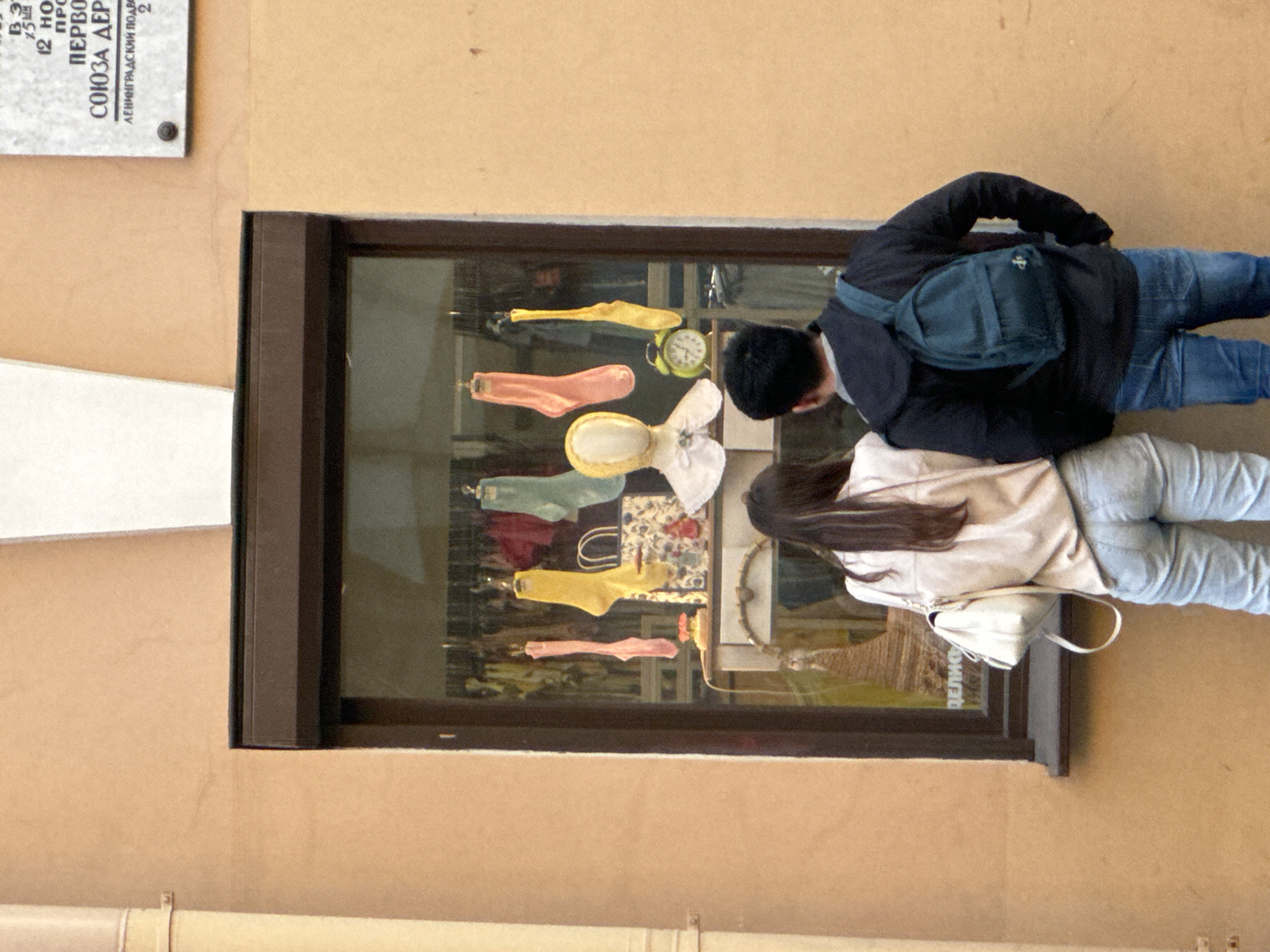 This is a medium shot, taken from a slightly elevated perspective, of a young couple standing in front of a storefront window on a street in Saint Petersburg, Russia. The couple, a woman on the left and a man on the right, are facing the window, both looking in. They are dressed in casual clothing; the woman wears a light-colored jacket and light-wash jeans, with a white purse slung over her shoulder. The man wears a dark hooded jacket with a dark green backpack and blue jeans.
The storefront window displays various items, including several brightly colored socks hanging from the ceiling, a mannequin head wearing a white collar, a decorative bag, and a yellow alarm clock. Behind these items, other goods are visible in the dimly lit shop. The building exterior is a warm, peachy-tan stucco. To the upper right of the window, a rectangular plaque with white lettering and dark text is affixed to the wall.
The street appears to be cobblestone, though this is only partially visible at the bottom of the frame. The overall impression is of a casual urban scene, possibly on a street with shops selling clothing or accessories.