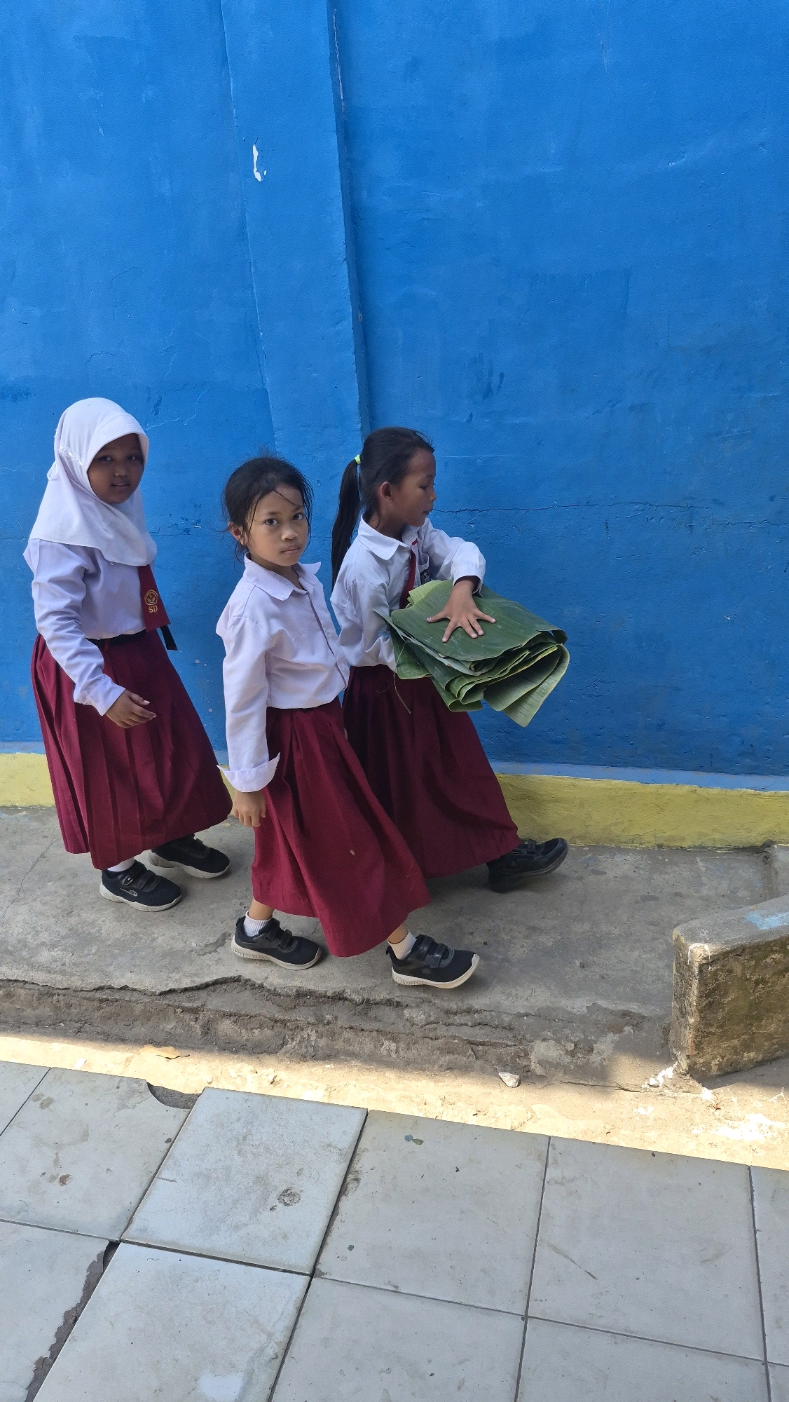Three young girls, dressed in white shirts and maroon skirts, walk along a pathway next to a bright blue wall in Putak, Indonesia. The girl on the left wears a light pink hijab. The middle girl, facing the camera, has dark hair pulled back. The girl on the right carries a stack of large, folded green leaves. Their shoes are dark and practical. The environment appears to be outdoors, with a concrete pathway and a textured blue wall that has some yellow trim near the ground. A concrete block is visible to the right. The lighting suggests it is daytime, with some shadows cast on the pathway.