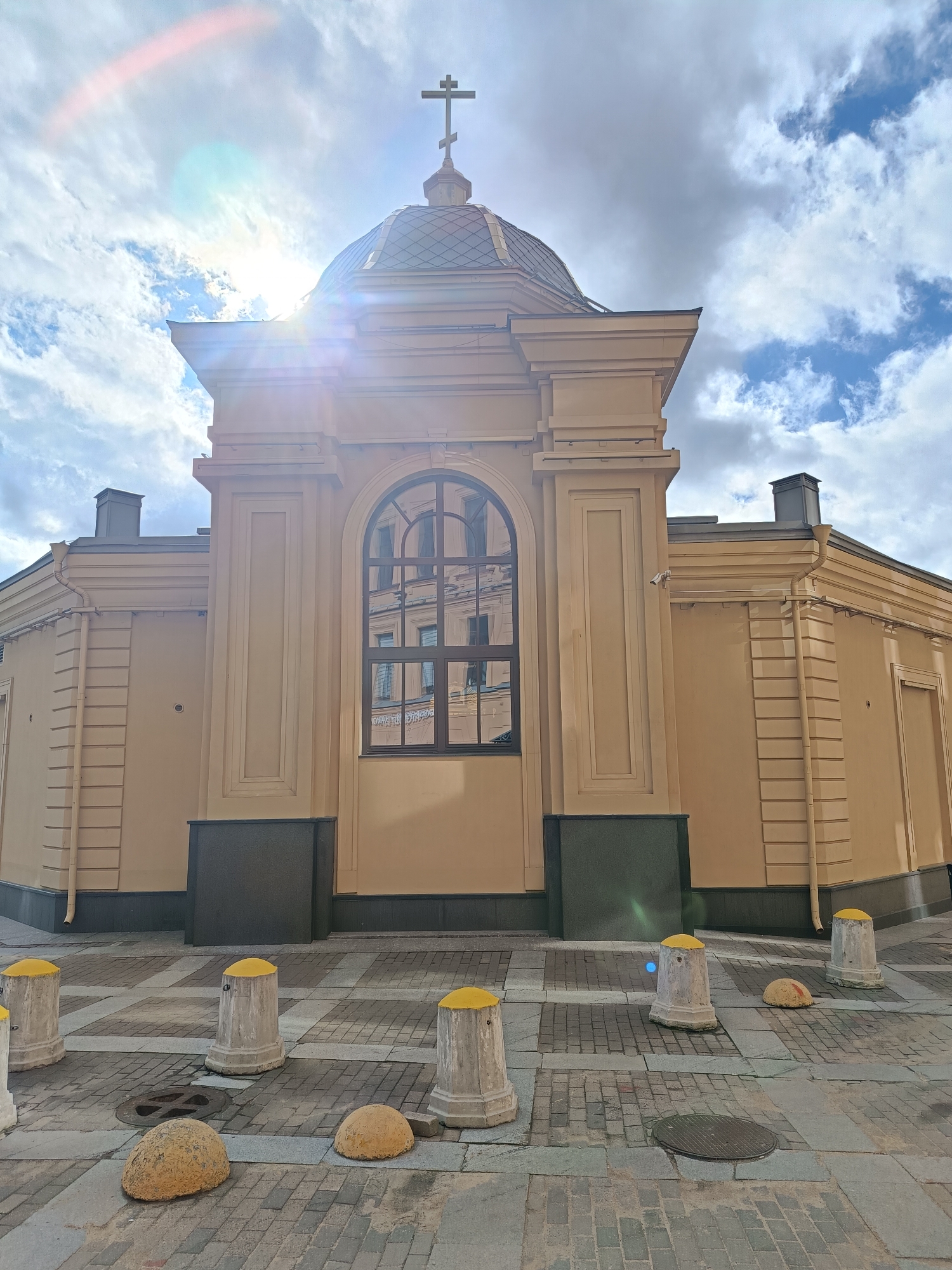 A low-angle shot of a building with a cross atop its dome, situated in an urban environment. The building is painted in a light, yellowish-tan color, and it features a large, arched window reflecting another building across the street. The architecture incorporates classical elements such as pillars and defined moldings. The base of the building is accented with dark, rectangular panels. In the foreground, a cobblestone or brick-paved area is populated with several short, concrete bollards, some topped with yellow caps. The sky above the building is partly cloudy, with bright sunlight causing lens flare, casting a dynamic play of light and shadow across the scene. The location is the city of Unknown, Unknown.