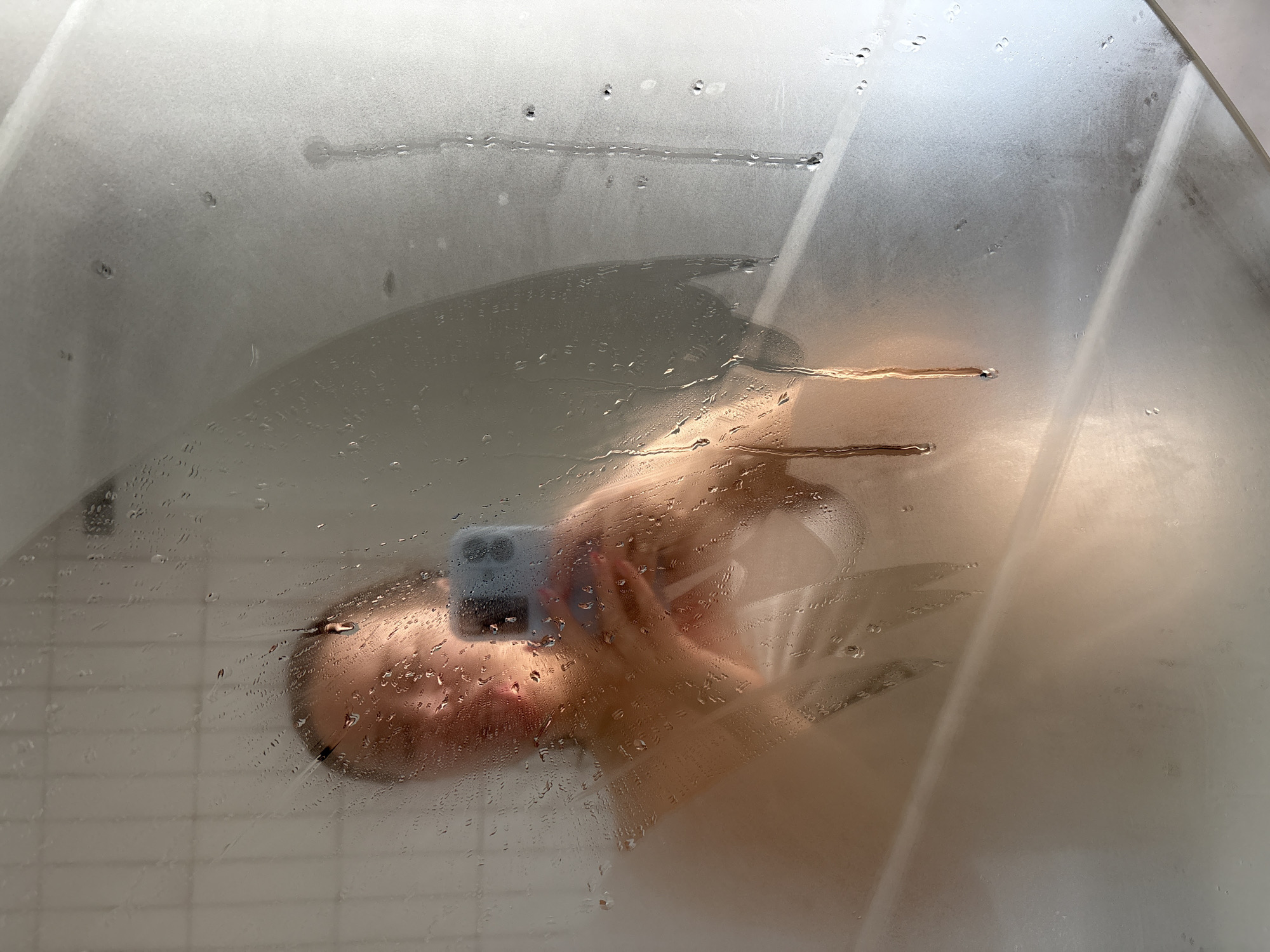A selfie is taken through a wet, frosted glass shower door. A person is holding a smartphone in their right hand, obscuring part of their face. The person has light skin. Drops of water cover the glass surface. The background suggests a tiled shower interior. There is no information to determine this is taken in Izhevsk, Russia.