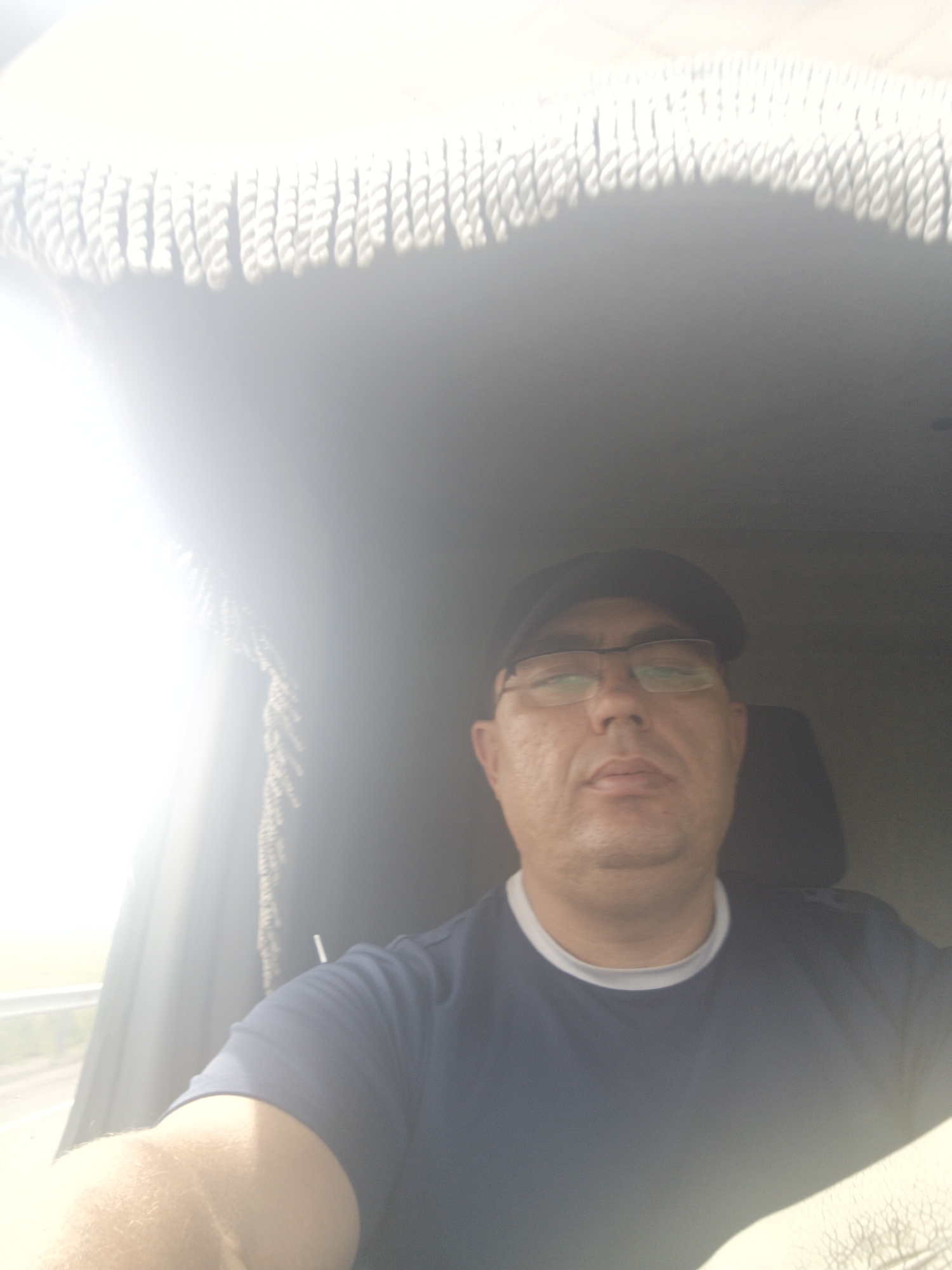 A selfie taken from the perspective of someone sitting inside a vehicle, likely a truck or van. The subject is a middle-aged man wearing a dark baseball cap and eyeglasses. He is looking directly at the camera with a neutral expression. The man is wearing a dark blue t-shirt with a lighter colored collar. The interior of the vehicle is visible, including a fringe-like decorative trim along the top of the windshield and a headrest of a seat behind him. Sunlight is streaming in from the left side of the frame, creating a bright glare and obscuring some details of the environment outside the vehicle. There is no information in the image to suggest the location in the city of Gusyne, Russia.