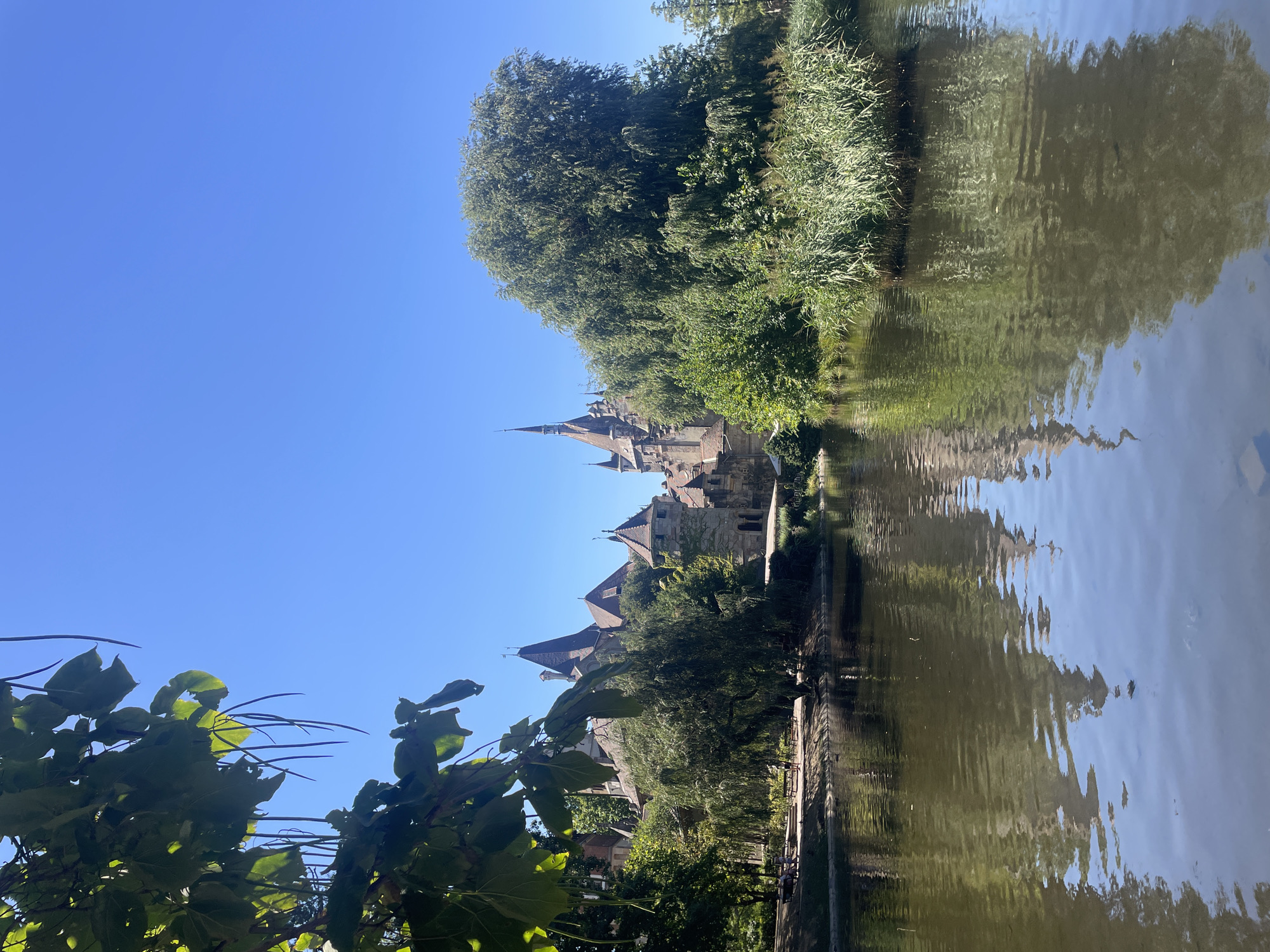 The image depicts a scenic view of Vajdahunyad Castle in Budapest, Hungary, on a clear, sunny day. A body of water, likely a lake or pond, occupies the foreground, with its surface reflecting the castle and surrounding greenery. The castle itself is a complex architectural structure with multiple towers, spires, and varying rooflines, constructed from stone and brick. Lush green trees and dense foliage flank the castle and line the water's edge. In the mid-ground, a pathway with benches and a few indistinct figures can be observed, suggesting a park or recreational area. The sky above is a vibrant, cloudless blue, indicating fair weather. The overall scene conveys a sense of tranquility and historical grandeur.