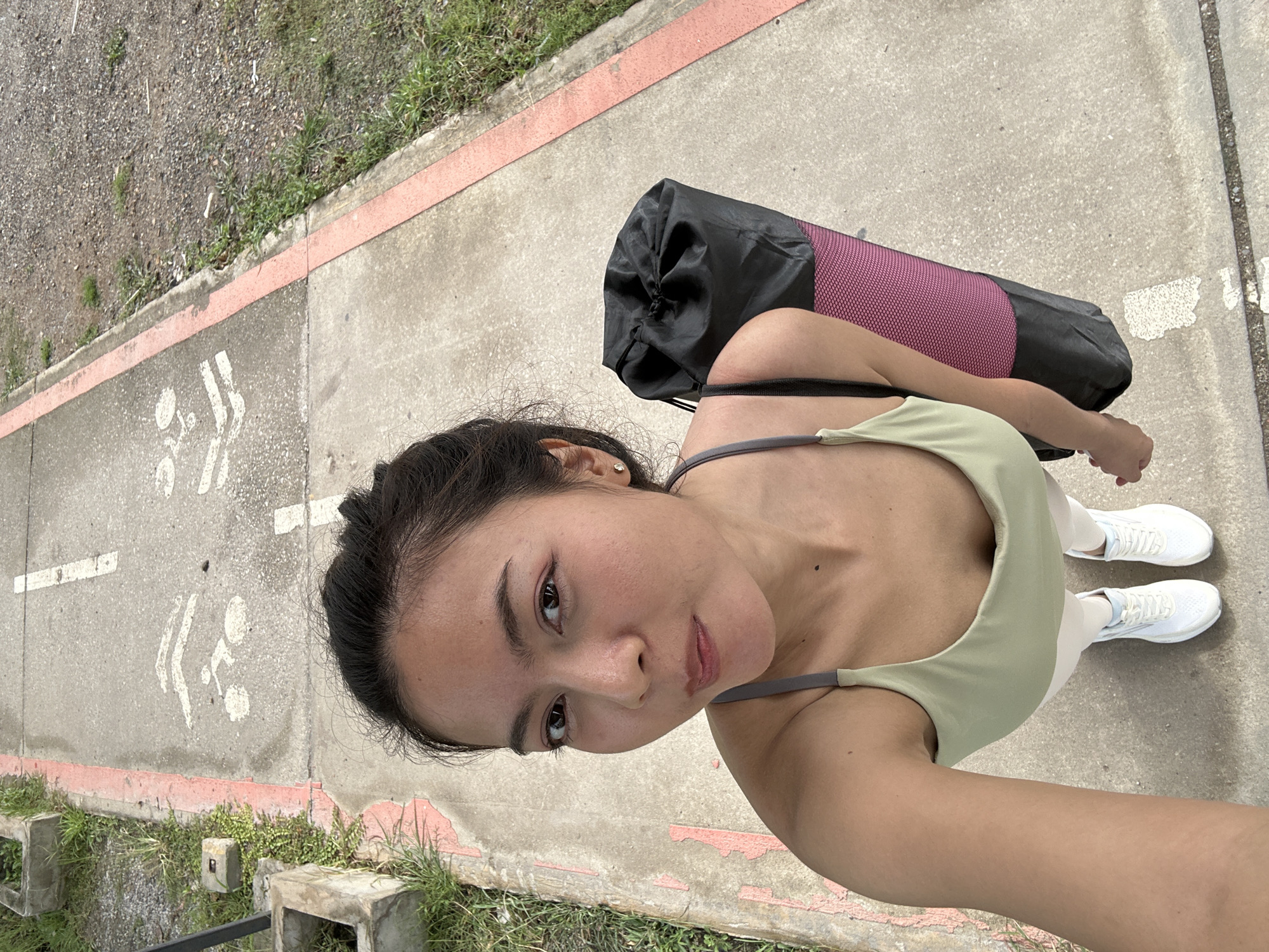 An Asian woman is taking a selfie outdoors. She is wearing a light green sports bra, white leggings, and white sneakers. She has her dark hair pulled back and is wearing small stud earrings. A black yoga mat bag with a pink mesh pattern is slung over her right shoulder. The woman is standing on a paved path that has painted bicycle lane markings. The path is bordered by a red stripe on the left and gravel and grass on the right. This scene is located in Tha Khlong Town Municipality, Thailand.