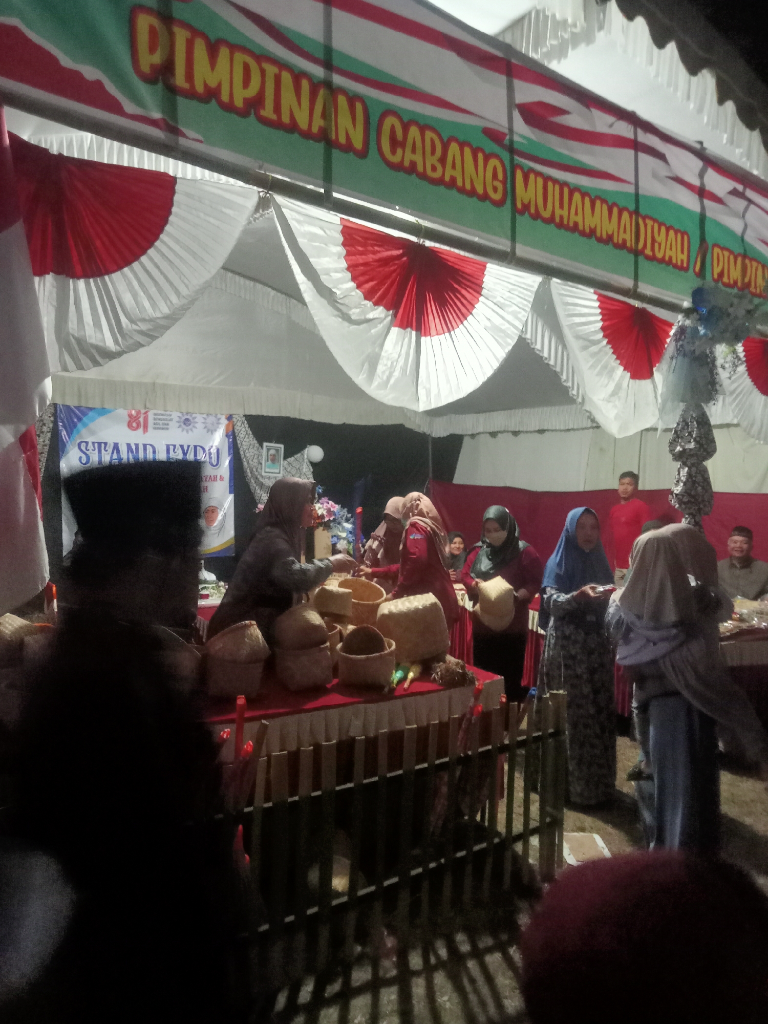 The image captures an outdoor event, likely a stand or bazaar, under a large tent adorned with red and white pleated fabric decorations. A prominent banner at the top reads "PIMPINAN CABANG MUHAMMADIYAH / PIMPIN." Below this, a smaller banner displays "81 STAND EXPO MUHAMMADIYAH & ..." with a graphic of a shield and a face.
Several people are visible in the scene. In the foreground, a person is out of focus, wearing dark clothing and a head covering. To the right of this person, a group of individuals are gathered around tables laden with woven baskets of various sizes. A woman in a brown hijab is seen holding a basket, and another woman in a blue hijab is looking down at something in her hands. Further back, a man in a red shirt stands near a tent pole, and to his right, a man in a brown shirt and a cap is seated at a table.
The environment appears to be an outdoor setting, with uneven ground visible and shadows cast by the tent and fencing. The overall lighting suggests it is either dusk or night, with artificial lighting illuminating the area. The presence of the banner with "MUHAMMADIYAH" suggests the event is organized by or associated with the Muhammadiyah organization, which is a major Islamic organization in Indonesia. The location is Pule, Indonesia, as indicated. The scene depicts a community gathering or a trade fair with elements of cultural display.