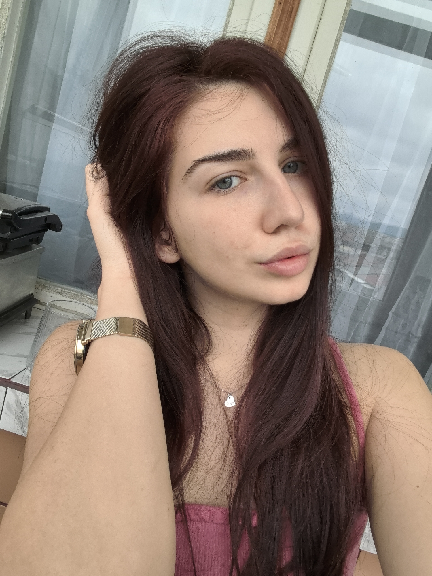 A young woman poses for a selfie indoors. She has long, dark auburn hair and blue eyes. She is wearing a pink top and a gold watch. Her left hand is raised, touching her hair. A silver necklace with a heart-shaped pendant is visible. The background shows a window with a view of a city, possibly Stara Zagora, Bulgaria, given the prompt's request. A stainless steel appliance sits on a marble or tile surface near the window. A white curtain partially covers the window.