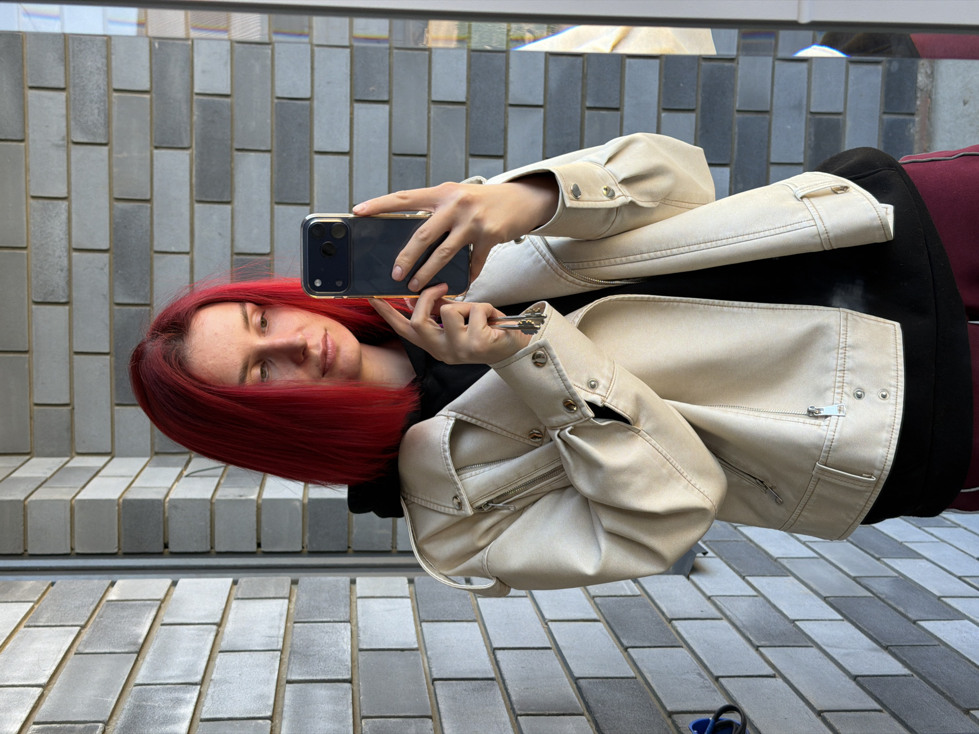 A woman with red hair is taking a selfie with a smartphone, holding keys in her other hand. She is wearing a beige leather jacket over a black hooded sweatshirt, with a burgundy garment partially visible underneath. The background consists of a wall with gray rectangular blocks, and a paved ground of similar color. The location is Chaikovsky, Russia, based on the gray blocks of the wall.
