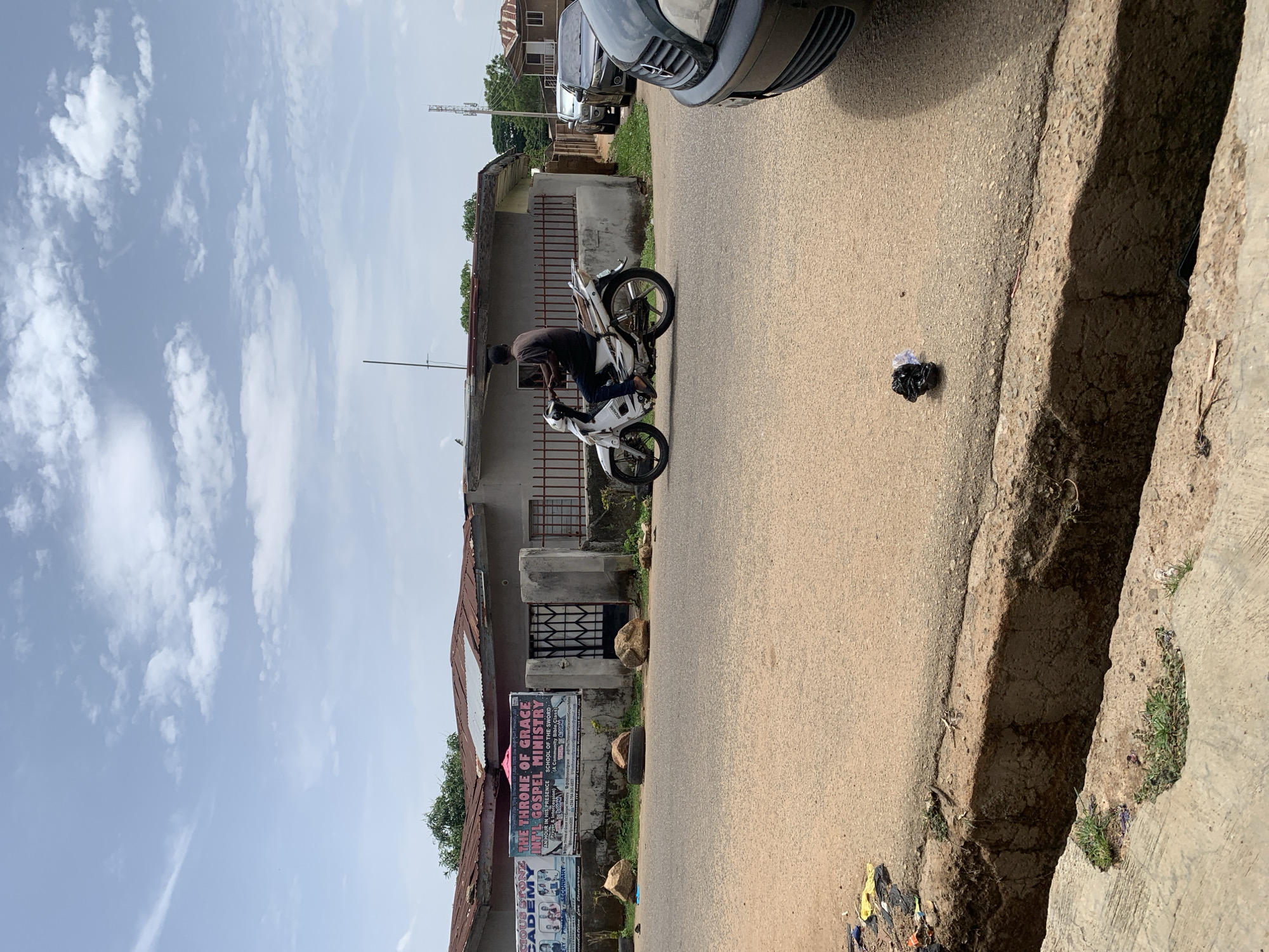 The image depicts a street scene in Jos, Nigeria. A person rides a white motorcycle on a paved road. The foreground includes a concrete curb and various debris. On the left, a wall displays several posters. One prominently features the text "THE THRONE OF GRACE INTL GOSPEL MINISTRY SCHOOL OF THE SWORD" and "AN HOUR IN HIS PRESENCE". Another poster mentions "CIOUS STONG CADEMY". A parked car is visible partially in the top of the frame. The sky is cloudy and the general environment appears to be a residential or commercial area.