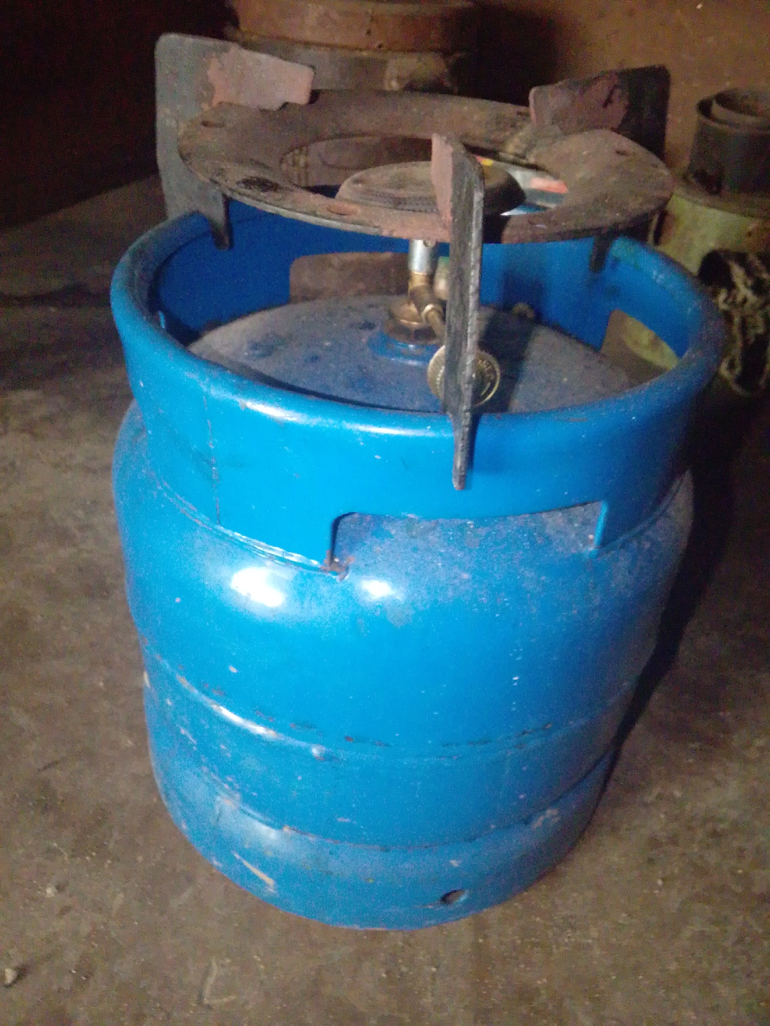 The image shows a blue gas cylinder with a cooking stove on top. The cylinder is cylindrical in shape and appears to be made of metal. The stove is a portable cooking appliance with a burner. The scene is indoors, with a rough, concrete floor. The lighting is dim, suggesting it is likely nighttime or an area with limited light. The background is dark and out of focus, but appears to show other objects and possibly part of a wall or structure. The overall impression is of a basic, functional cooking setup. The location is specified as Jalingo, Nigeria.