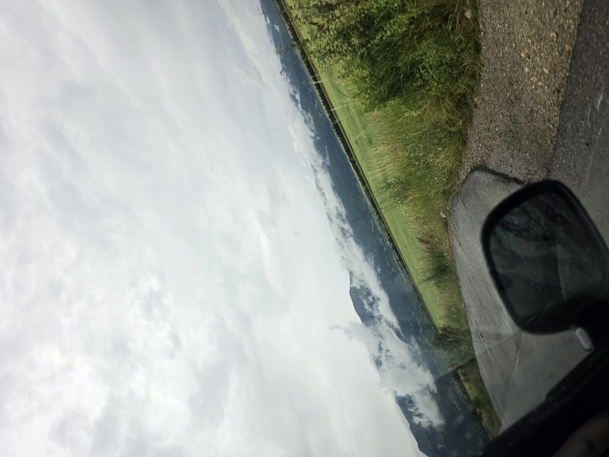 A tilted, eye-level, outdoor, daytime, full shot captures a view from a vehicle traveling along a paved road towards a lush green field and misty mountains. The road, wet and appearing to be asphalt with some gravel on the shoulder, curves slightly to the right. In the lower right corner, a black side-view mirror is visible, reflecting the overcast sky. Beyond the road, a vibrant green field stretches towards a range of dark green mountains partially obscured by wisps of white clouds. Tall, dry grass and green bushes line the edge of the field and road. The sky is filled with a dense layer of white and gray clouds, suggesting a potentially overcast or rainy day. The location is identified as being in Manolovo, Bulgaria.