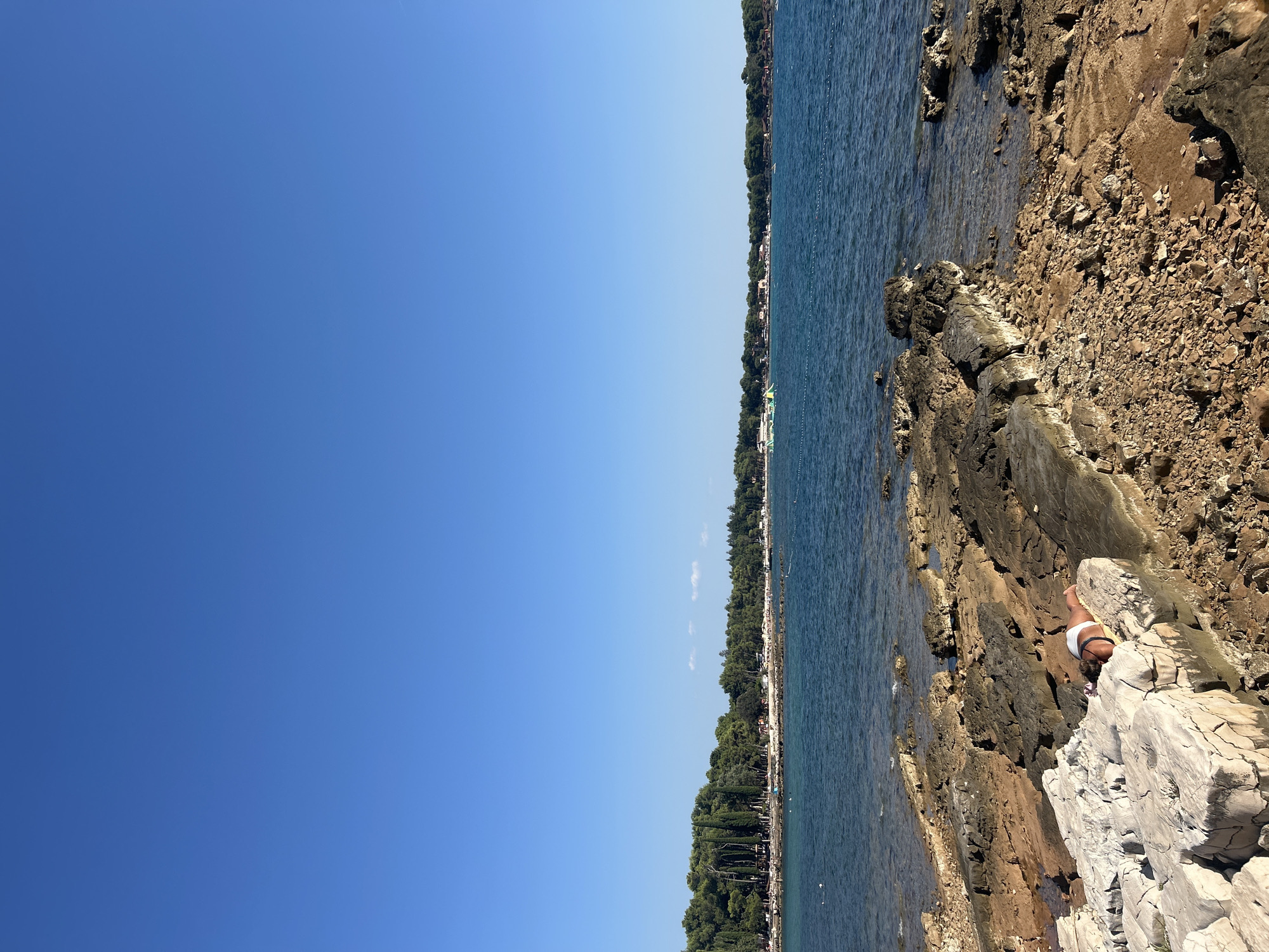 The image shows a rocky coastline with the blue sea and sky. In the foreground, a person is lying on their stomach on a large white rock, sunbathing. The person is wearing white bikini bottoms. The rocky shore extends into the clear blue water. In the background, there is a dense line of green trees, with a few buildings visible behind them. Some boats are in the water further out. The scene appears to be a popular tourist spot, possibly Katoro, Croatia, given the clear blue skies and the presence of a beach and boats. The overall impression is one of a sunny, relaxing day at the seaside.