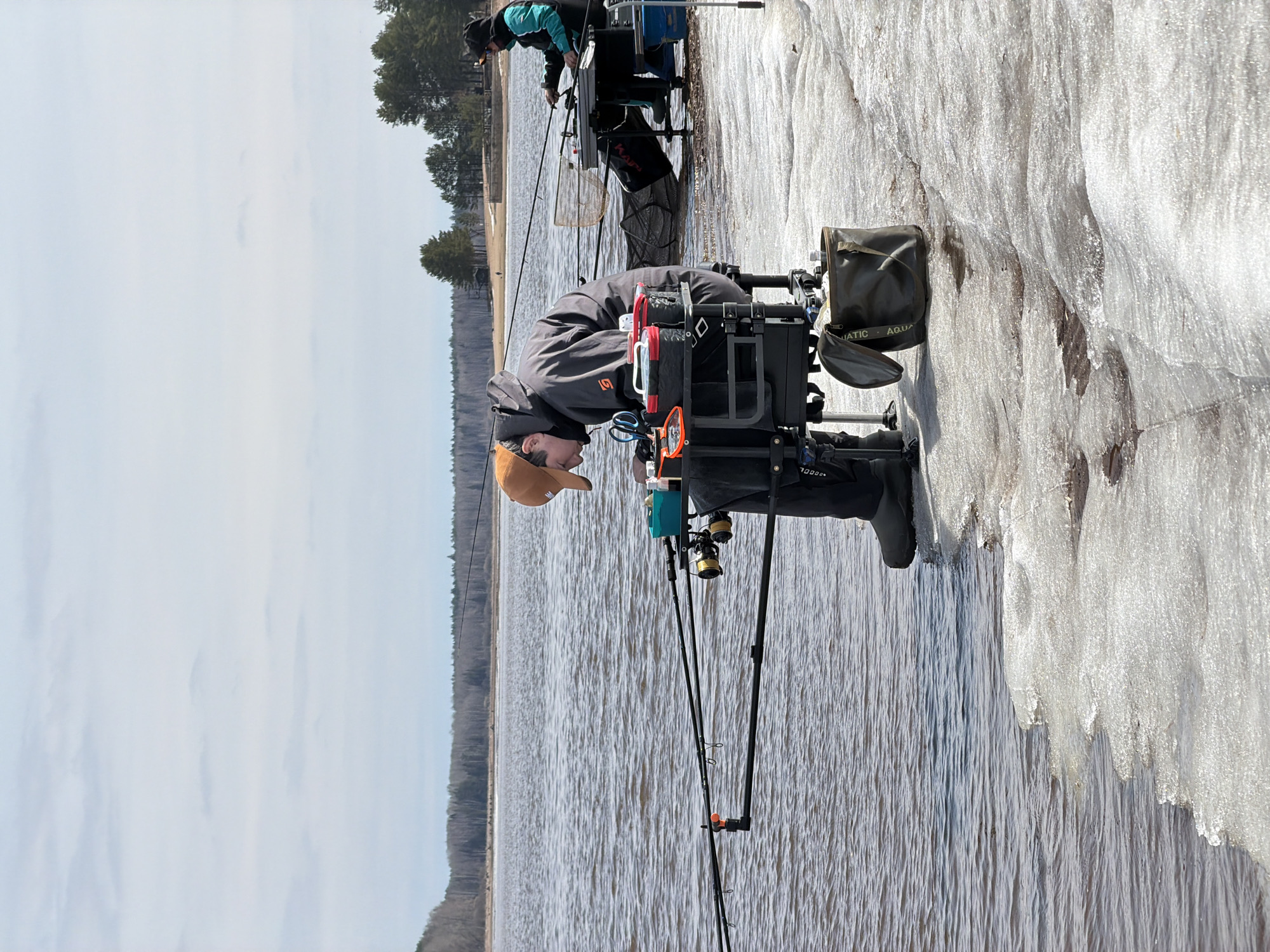 This media file captures an outdoor fishing scene on a bright, cold day. Two individuals are engaged in feeder fishing from a partially frozen bank.
The closest person, in the lower-center of the image, is seated on a specialized fishing station or seat box. They wear a grey jacket with an "S5" logo on the sleeve, dark trousers, black boots, and an orange baseball cap. Their fishing rods are set up in front of them, extending towards the water, and their station is equipped with multiple accessories, including a green "AQUATIC" branded bag and what appears to be a bait bucket. "7000xx" is visible on part of their fishing chair setup.
Further back, towards the upper-center, a second angler is similarly positioned, wearing a teal and black jacket. Their fishing station also has rods cast into the water, and a black and red bag with "Karp" written on it, along with a keepnet, is visible near their setup.
The setting depicts a body of water, possibly a river or lake, on the left side of the image. The surface of the water shows ripples. The right side of the image is dominated by a substantial amount of ice or hardened snow covering the bank where the anglers are situated. In the background, beyond the water, a distant tree line of dark coniferous trees is visible under a pale, slightly overcast sky.
The scene suggests early spring or late winter conditions in a northern climate, consistent with the provided location context of Chaikovsky, Russia. The anglers appear focused on their activity.