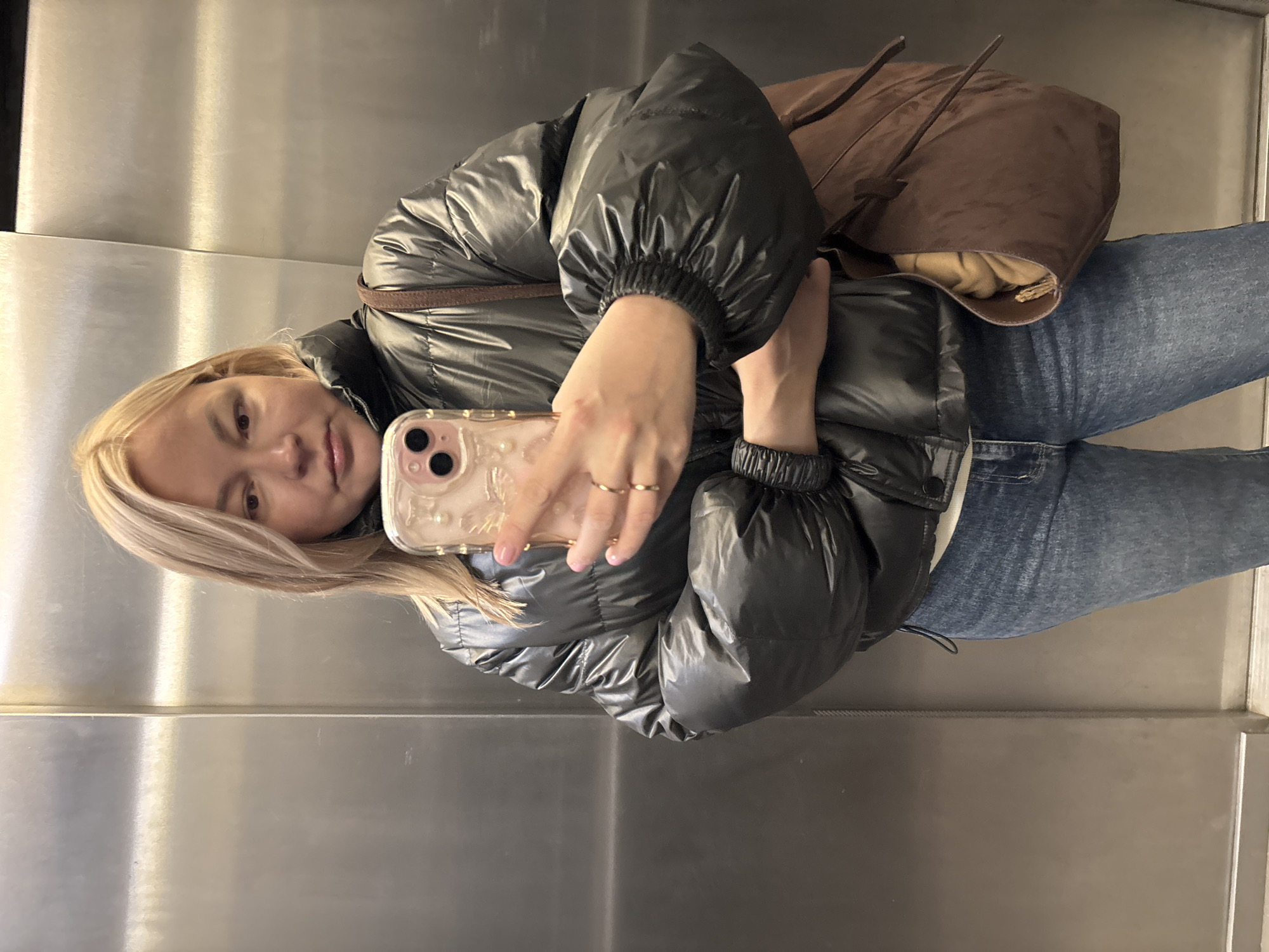 A fair-skinned woman with blonde hair is taking a selfie with her smartphone in what appears to be an elevator. She is wearing a shiny dark puffer jacket, blue jeans, and has a brown bag slung over her shoulder. Her phone has a light-colored case. The elevator has brushed metal walls. There is no evidence to indicate this photo was taken in Izhevsk, Russia.