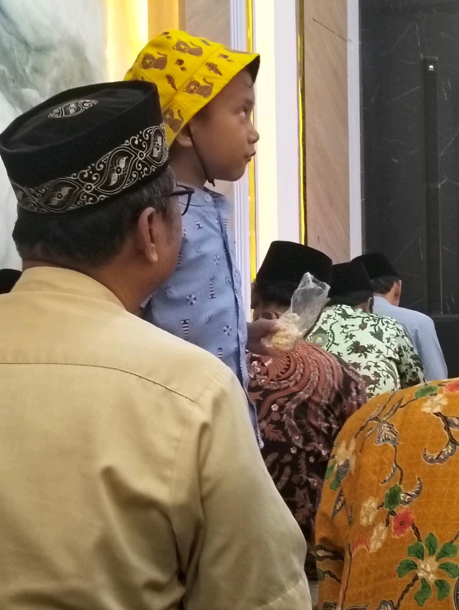 The image shows a group of people, including a young boy wearing a yellow hat with yellow bear prints, a man in a black cap with silver embroidery, and others wearing traditional Indonesian attire. The setting appears to be an indoor gathering, possibly a religious event, given the attire and headwear. The background features a wall with textured patterns and vertical dark panels. The people are seen from the back and side, with varying levels of focus. The composition suggests a candid or observational style of photography, capturing a moment within a community. The specific location in Dagan, Indonesia, is indicated.