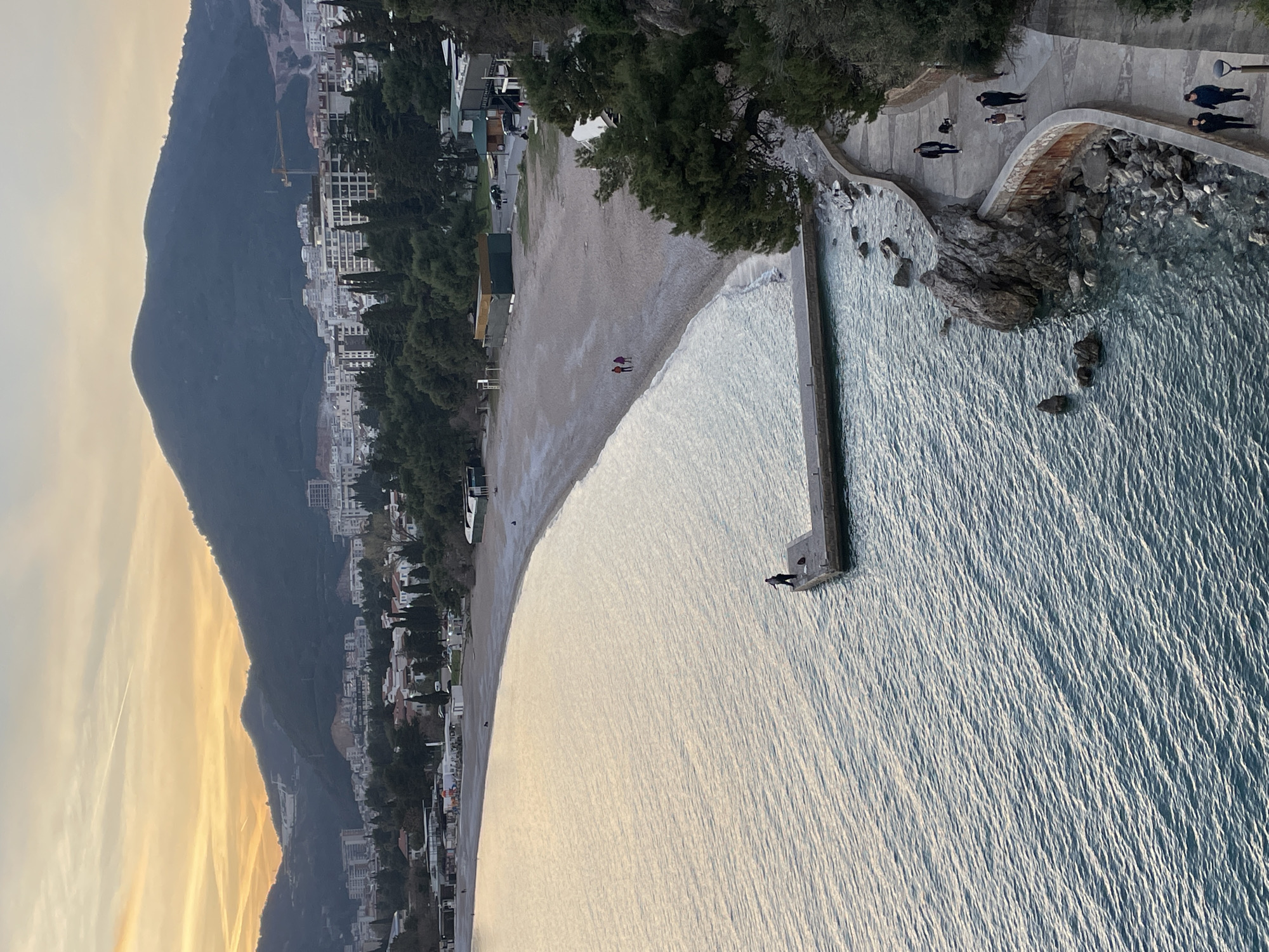 The image depicts a panoramic view of the coastal area in Budva, Montenegro, observed during what appears to be late afternoon or early evening due to the warm hues in the sky. The foreground is dominated by calm, rippled blue-green water.
To the right, a concrete promenade curves along a rocky coastline, with several individuals walking on it. One person walks a dark-colored dog. Further along the promenade, two distinct pairs of individuals are observed walking. The rocky shoreline beneath the promenade features scattered rocks and sparse vegetation.
Extending into the water from a sandy-pebbly beach is a grey concrete pier. A solitary individual stands at the end of this pier, positioned with their back to the viewer, appearing to be fishing.
The beach itself forms a wide, crescent-shaped expanse of light-colored sand or pebbles. Two individuals in dark clothing are walking near the waterline on the beach. Behind the beach, a strip of green foliage separates it from several small, low-rise buildings, possibly commercial establishments or beach facilities. Vehicles are visible on a road behind these structures.
In the mid-ground, the city of Budva unfolds with numerous multi-story buildings, predominantly white or light-colored, interspersed with green trees. A large construction crane is visible in the distance among the buildings, indicating ongoing development.
The background features prominent, dark-green forested mountains that rise behind the city. The sky above the mountains exhibits soft orange and yellow tones, characteristic of either sunrise or sunset, with faint cloud patterns.