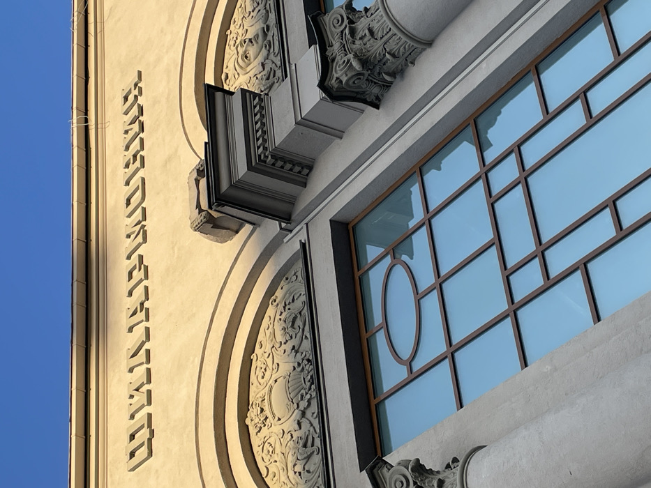 An upward-angled exterior view captures the ornate facade of the former Circus-Variety "Armoniya" building, now the Sverdlovsk Academic Drama Theatre, located at Prospekt Lenina, 48 in Yekaterinburg, Russia. The building's upper section, rendered in a cream-tan stucco, is bathed in direct sunlight from the upper left, creating distinct shadows. Raised Cyrillic letters across the top frieze spell "ЦИРКЪ" (Circus) and "АРМОНИЯ" (Armoniya) in pre-1918 orthography. Below this text, two arched recesses are visible, each containing intricate bas-reliefs. The left bas-relief depicts a central urn or basket flanked by stylized foliage and figures. The right bas-relief features a classical female face surrounded by leaves. Decorative architectural elements, including ornate cornices, sections of fluted pilasters, and Corinthian-style column capitals, frame these features. A large, multi-paned window occupies the lower portion of the frame, set within a brown grid-like frame, and features a prominent oval-shaped pane at its top center. The window glass reflects a clear, deep blue sky. The sky is also visible directly above the building, devoid of clouds. No people, actions, or interactions are present in the scene.