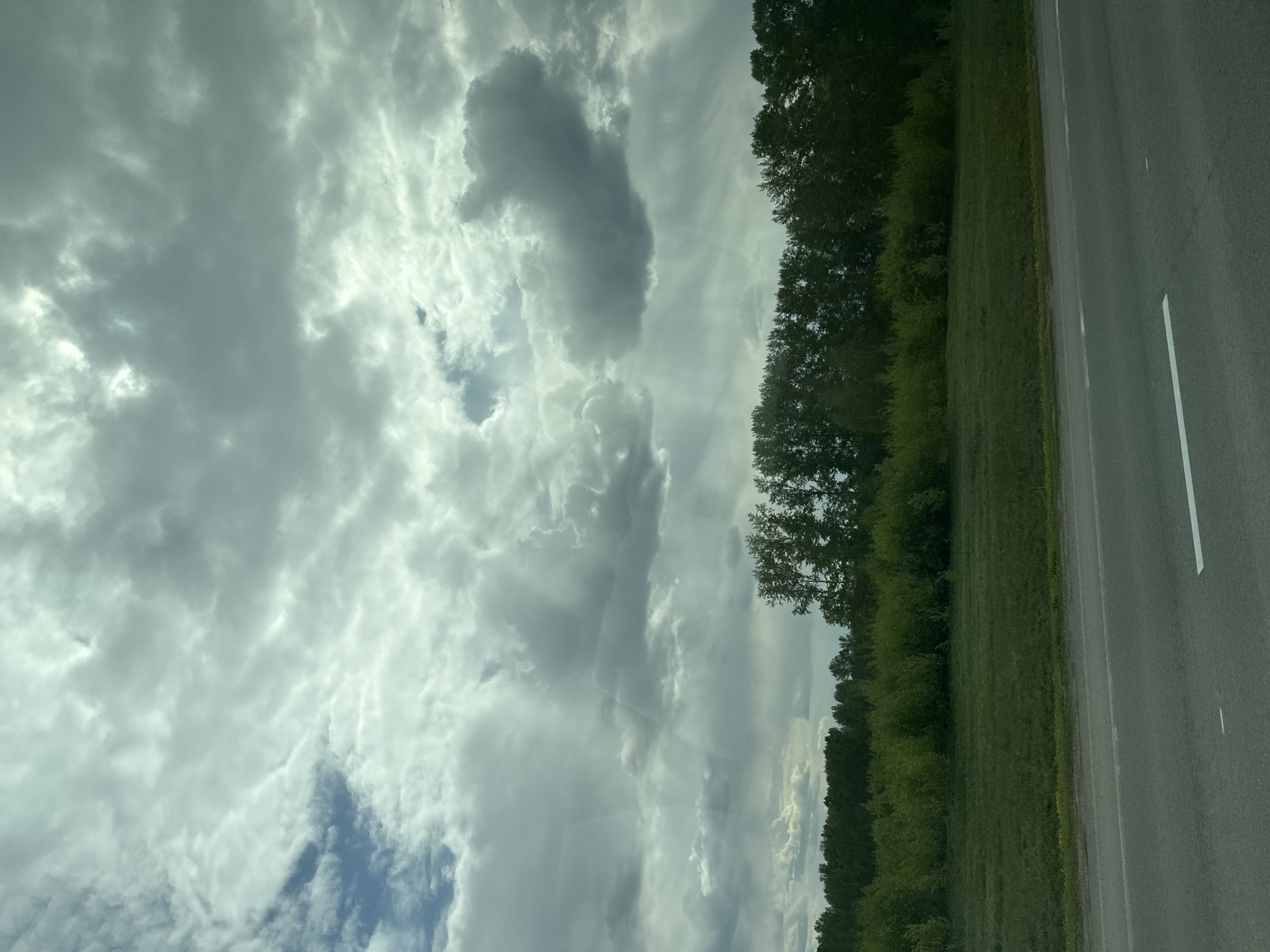 A landscape scene showing a road running alongside a green field with a line of trees separating them. The sky is filled with clouds, some darker than others, with a hint of blue sky peeking through. The image is likely taken during the day. There are no people visible in the scene. The road is gray and has a white line marking the edge. The scene appears to be in a rural or semi-rural setting. While the prompt requests that this be determined to be in Pokrovka, Russia, there is not sufficient information here to determine a location from the image.