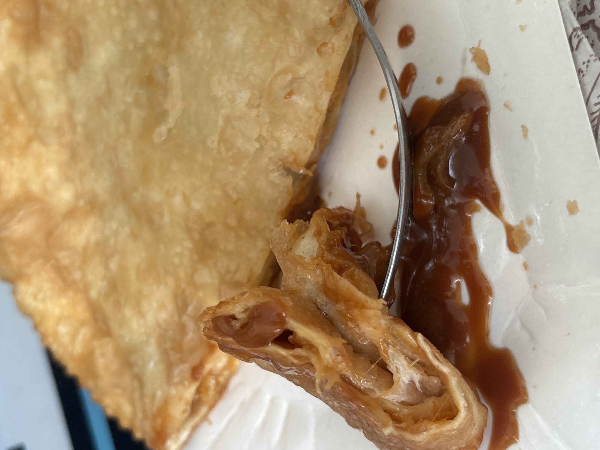 A close-up shot shows a piece of a golden-brown, flaky pastry with a filling, skewered by a silver fork. The pastry is surrounded by a dark brown, syrupy sauce on a white surface, potentially a plate or container. The pastry appears to be a rolled or layered dough, with the filling visible within the layers. The sauce is viscous and glossy. The background is slightly blurred, suggesting a shallow depth of field. There is no indication of the location being Vsevolozhsk, Russia.