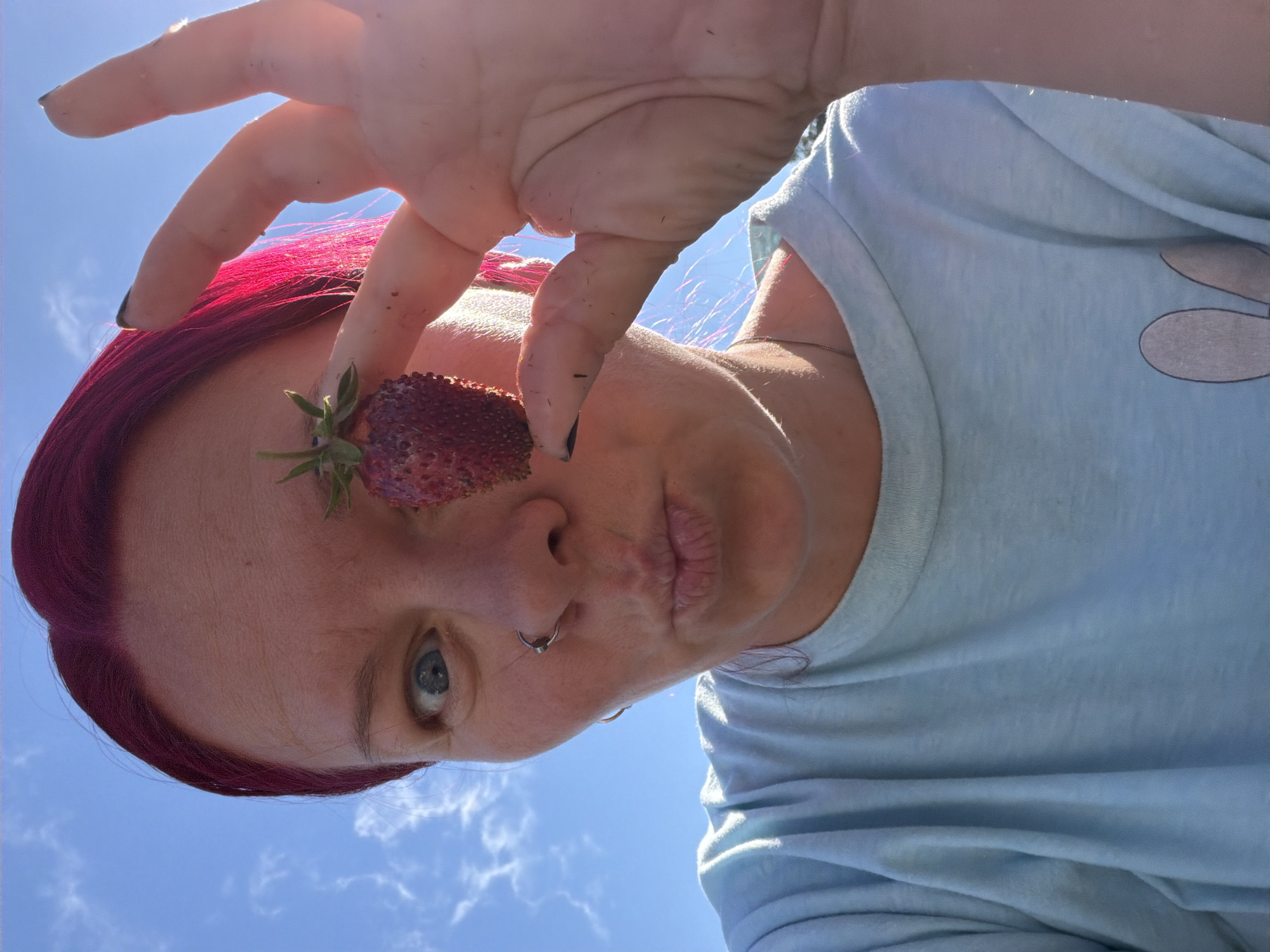 A woman with bright pink hair and a nose ring poses for a selfie, holding a single strawberry up to her eye. She is wearing a light blue t-shirt, and a silver necklace is visible. The background is a clear blue sky with some wispy white clouds, suggesting an outdoor setting. The sun is shining brightly, creating lens flare on the right side of the image. The woman is making a kissing face. The location is unspecified by any visual cues.