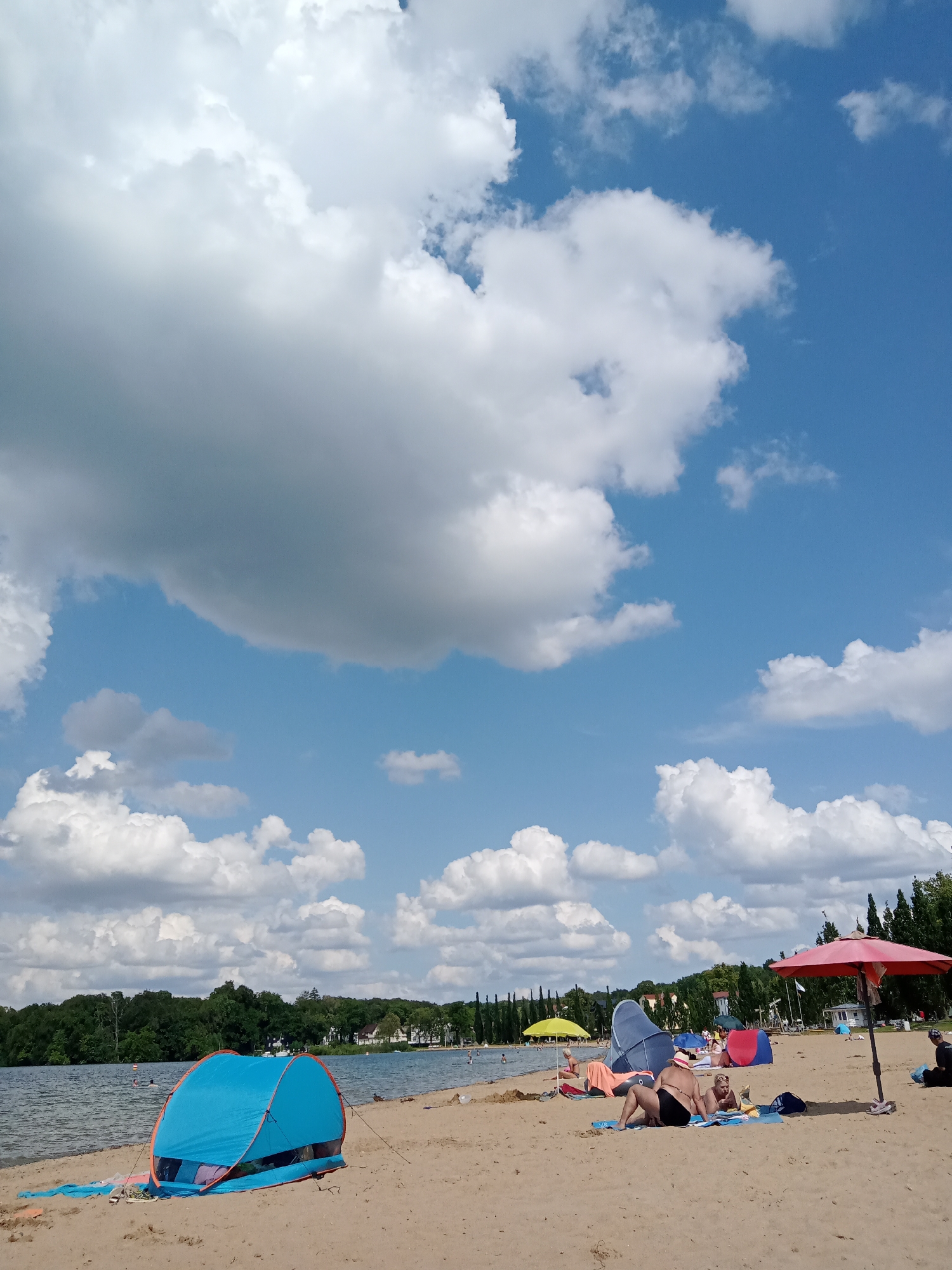 The image depicts a sunny day at a beach located on the shore of the Schweriner See in Schwerin, Germany. The foreground is dominated by a sandy beach with several beachgoers engaged in leisure activities. A bright blue pop-up tent with orange trim is set up on the left side of the frame, with a blue towel partially visible inside. Several people are sunbathing on towels, including a woman in a red bikini top and black swimsuit bottoms, and a child. A large red umbrella provides shade for another area of the beach. In the background, the calm waters of the lake stretch towards a tree-lined shore. Buildings are visible among the trees, suggesting a suburban or park area. Scattered white and grey clouds are present in a vibrant blue sky. The overall scene is one of a relaxed summer day at a popular recreational spot.