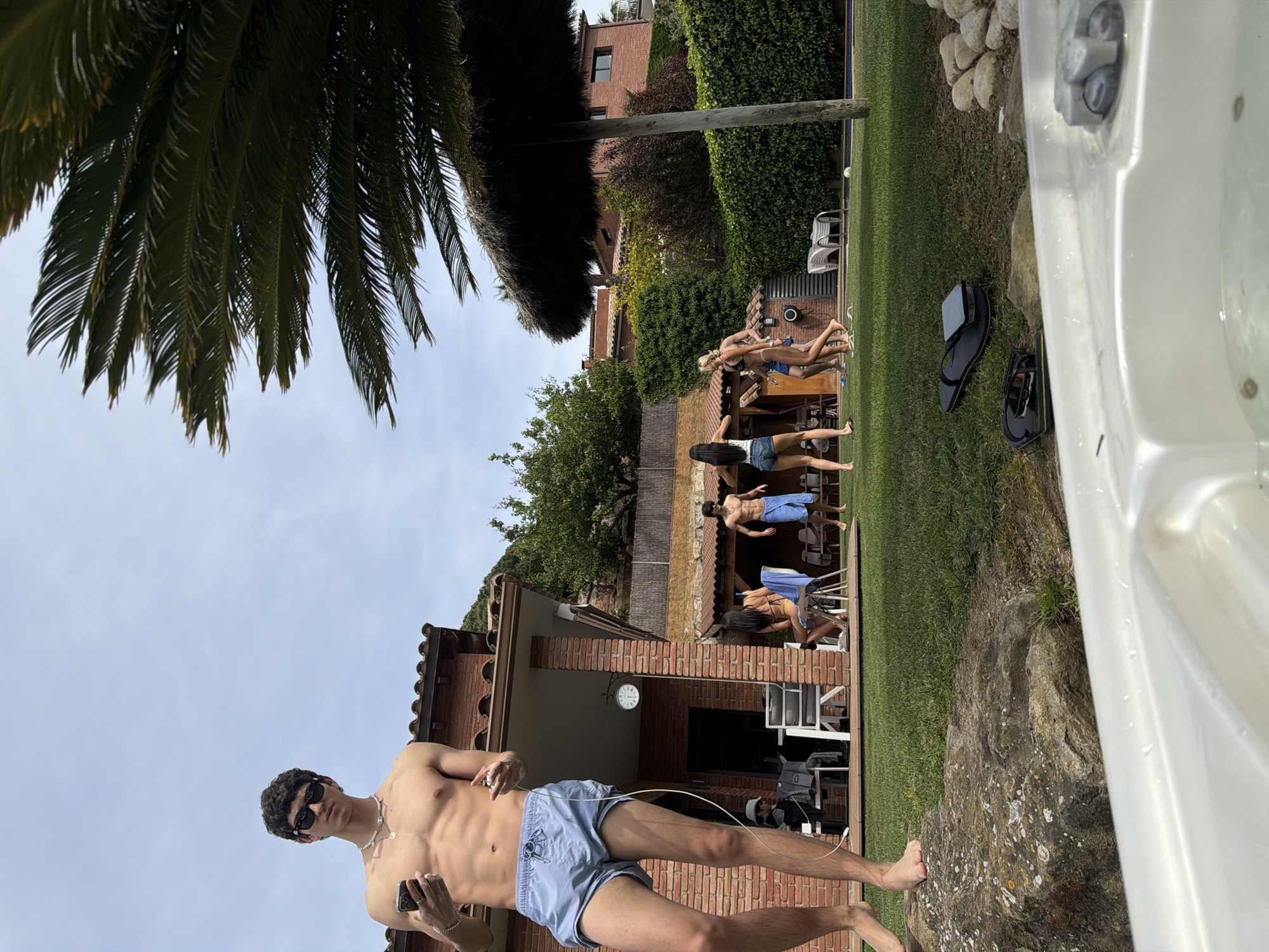 A shirtless young man in blue swim trunks, sunglasses, and earphones stands in the foreground of a backyard scene in Sant Andreu de Llavaneres, Spain. He holds a smartphone in his left hand. Behind him is a low brick building with a clock, and a group of people are sitting in chairs on the lawn near a pool. A palm tree hangs over the left side of the frame. A white hot tub is visible in the lower right corner. A thatched shade structure and other buildings are visible in the background.