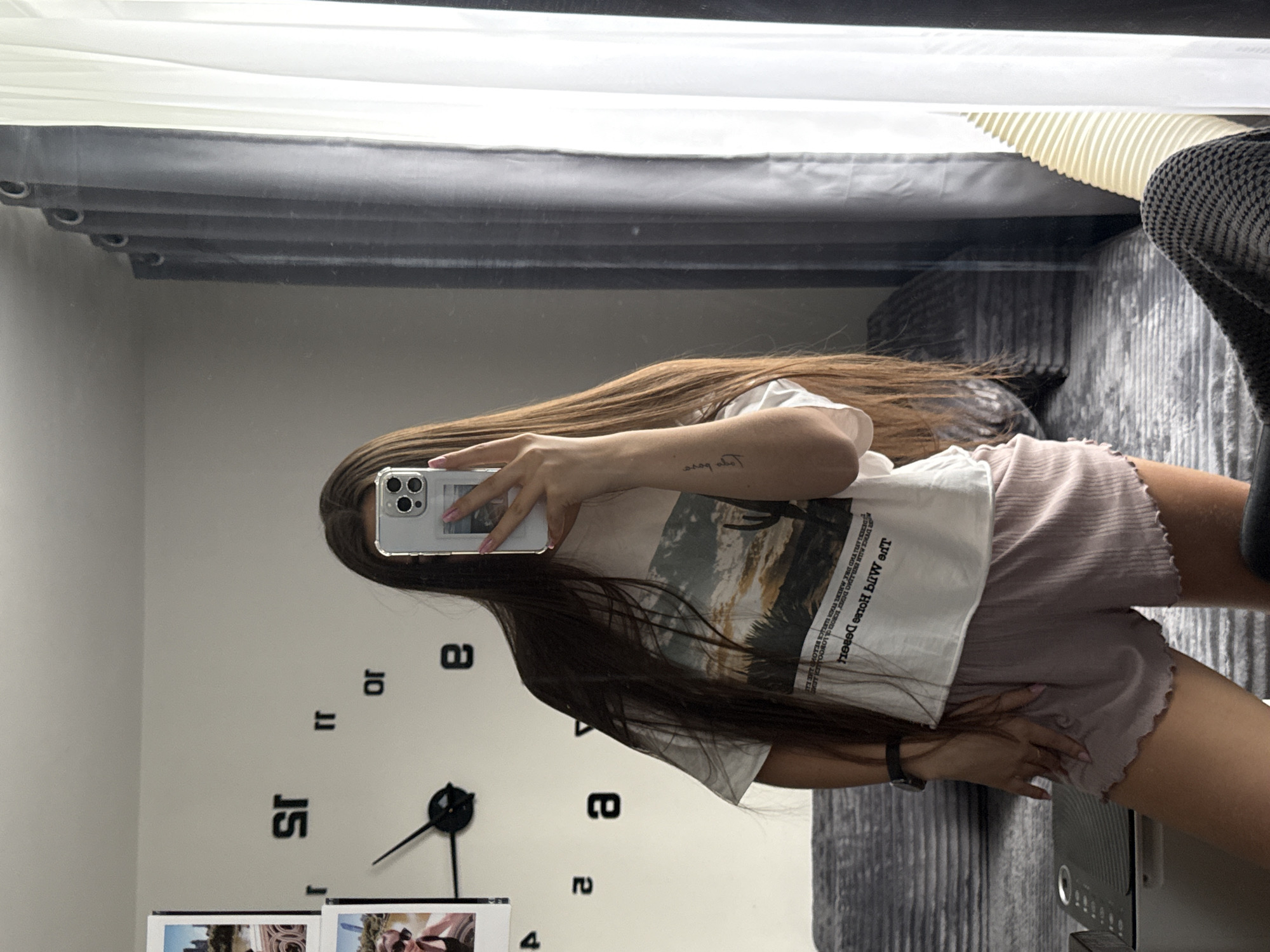 A young woman with long, brown hair is standing in front of a mirror, taking a selfie with her phone. She is wearing a white t-shirt with a desert landscape graphic and pink ruffled shorts. A large, modern clock with Roman numeral stickers is visible on the wall behind her. To her left, a framed photo of a city skyline is attached to the wall. The background includes a sofa and curtains, suggesting an indoor setting, possibly a bedroom or living room. The woman has a tattoo on her left forearm. The overall scene appears to be a casual, home environment. The image does not contain any elements that suggest a specific location in New York City, United States of America.