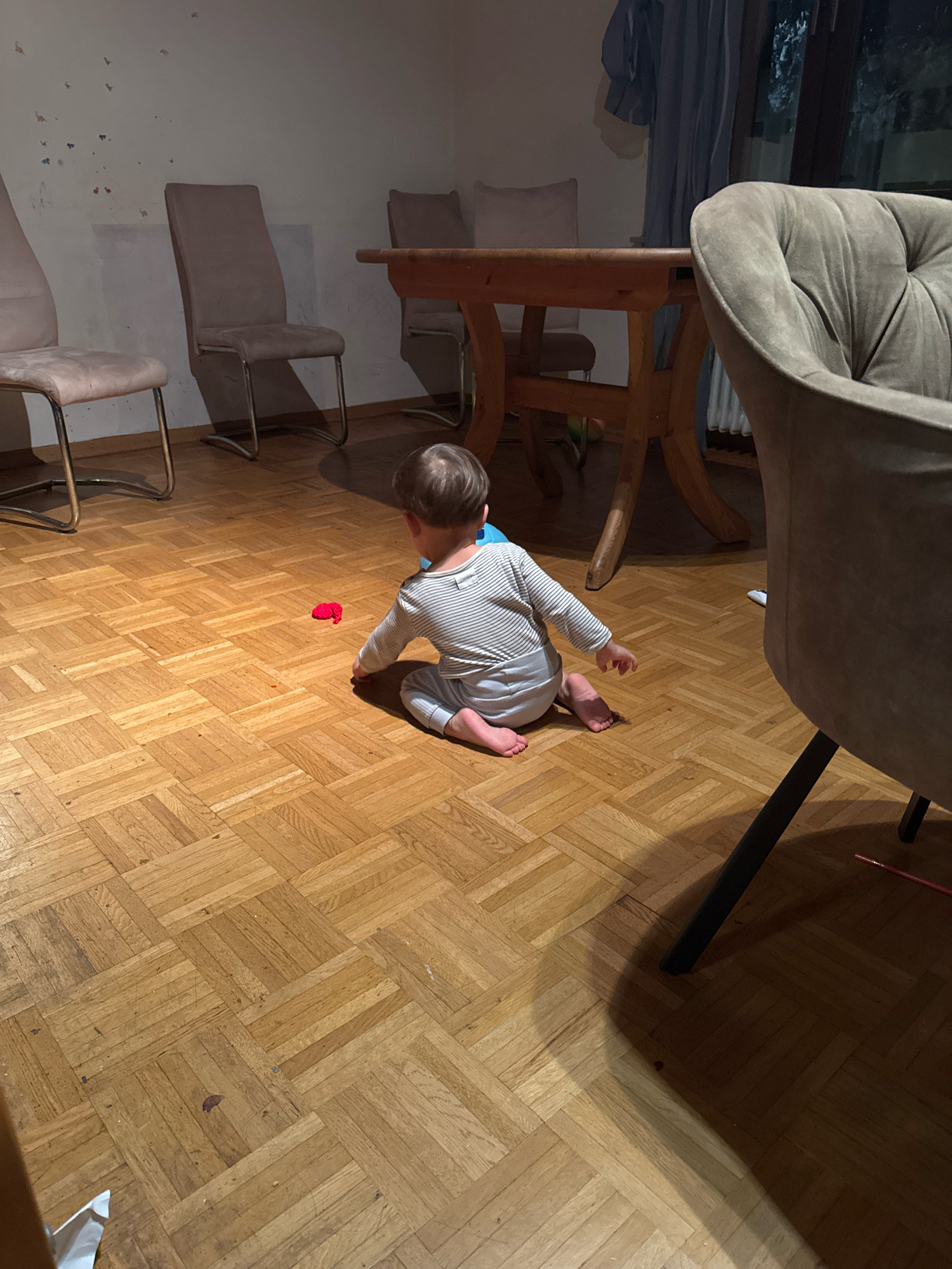 A toddler, dressed in a striped long-sleeved shirt and grey pants, sits on a wooden parquet floor in a dimly lit room. The toddler is facing away from the camera, looking towards a small red object on the floor. In the background, there is a wooden dining table with chairs and a large upholstered armchair. The room appears to be a dining area in a home in Überherrn, Germany.