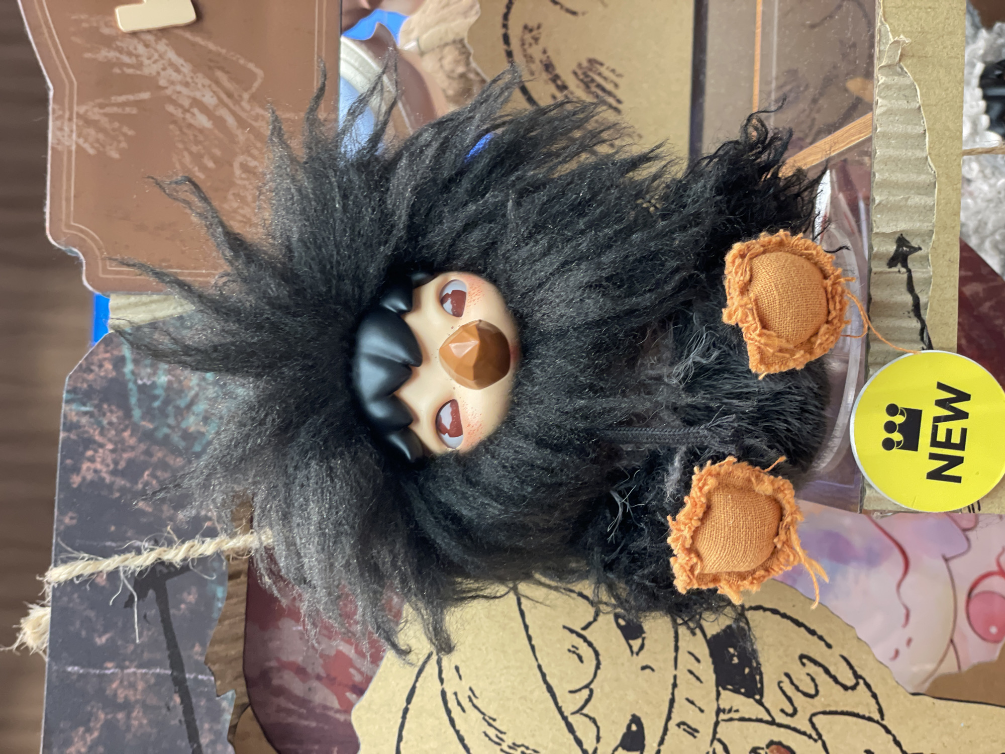 A black, fluffy, toy creature with a brown beak and large red eyes is the central focus of the image. The toy has orange fabric feet and a messy, spiky black head of hair. It is positioned in front of a cardboard background with a drawing of a creature with large eyes and a long, wavy mouth. A yellow circular sticker with a crown and the word "NEW" in black text is visible in the lower foreground. The scene appears to be a retail display, possibly in a store in Pattaya City, Thailand.