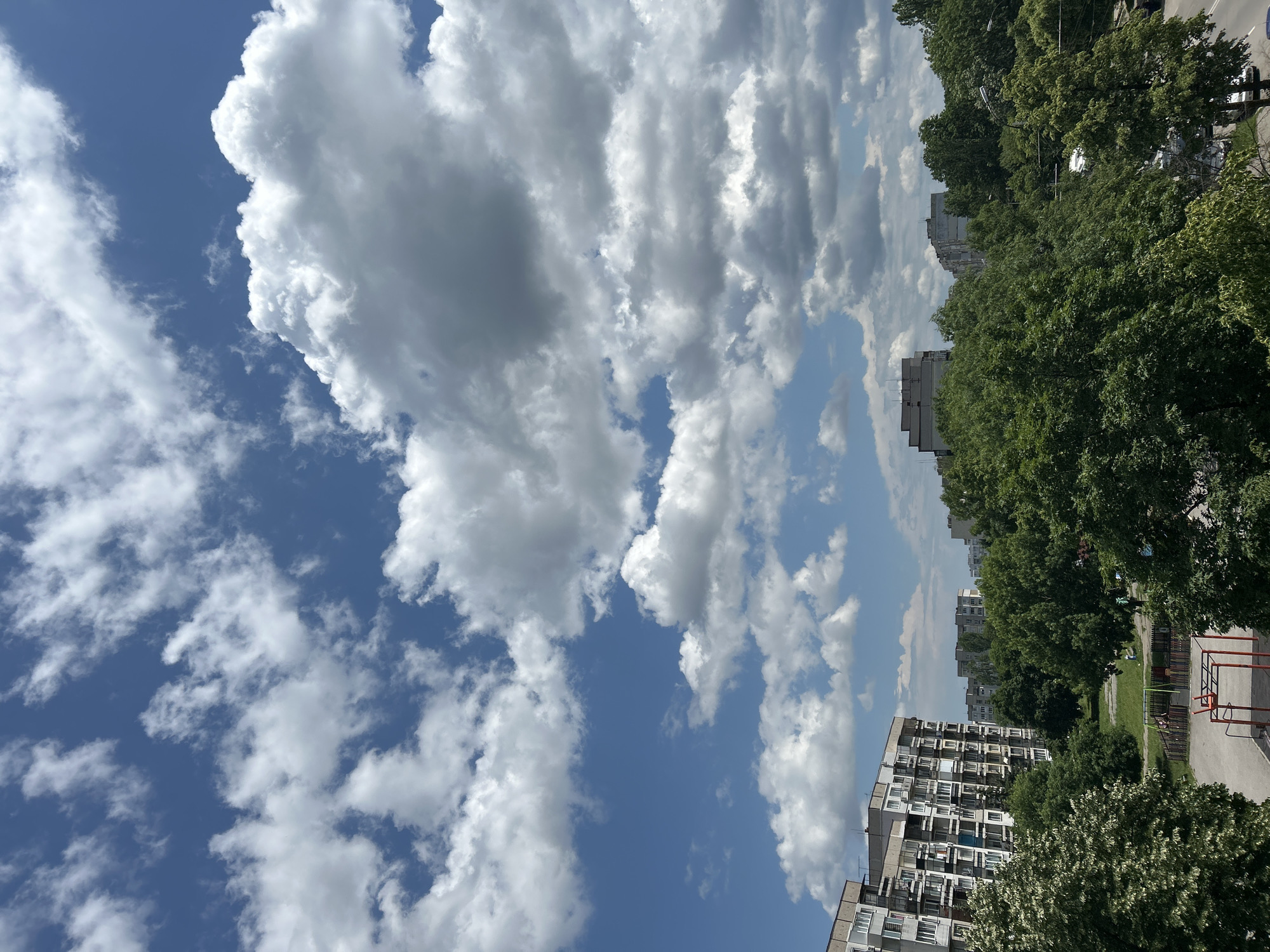 The image displays an elevated view of a residential area in Sofia, Bulgaria, under a bright, partly cloudy blue sky, characteristic of a daytime scene in late spring or early summer. The upper portion of the frame features a clear blue sky with scattered cumulus clouds of varying sizes and densities, ranging from bright white to light grey. The lower and mid-ground are dominated by dense green foliage of mature deciduous trees, some exhibiting light-colored blossoms.
In the left mid-ground, a prominent multi-story apartment building constructed from concrete panels, consistent with post-socialist architectural styles common in Eastern Europe, features numerous windows and balconies. Additional similar apartment blocks are visible in the background, partially obscured by the extensive tree canopy. Below the main apartment building, a public recreation area is visible, containing a children's playground with a red metal climbing structure and swings partially obscured by a fence. This area includes paved surfaces and grassy sections. No human figures are discernible within the frame. A roadway with parked vehicles and utility poles is partially visible through the trees on the right side of the image.