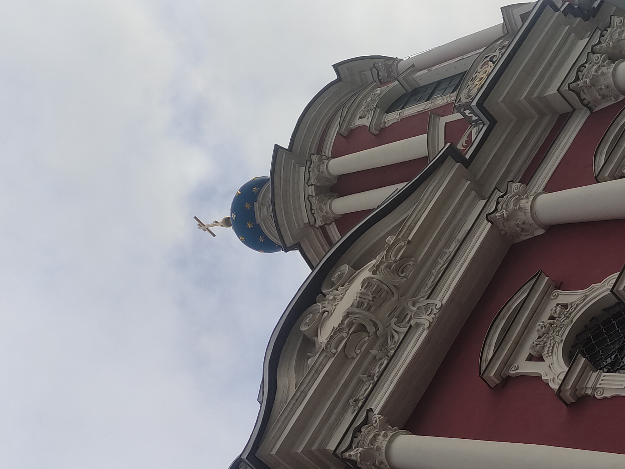 The image shows a low-angle view of a building in Moscow, Russia, likely a church. The architecture is ornate, with red walls, white columns, and elaborate decorative trim. A blue dome with gold stars is topped with a gold cross, indicating a religious building. The sky is overcast. There are no people visible in the shot.