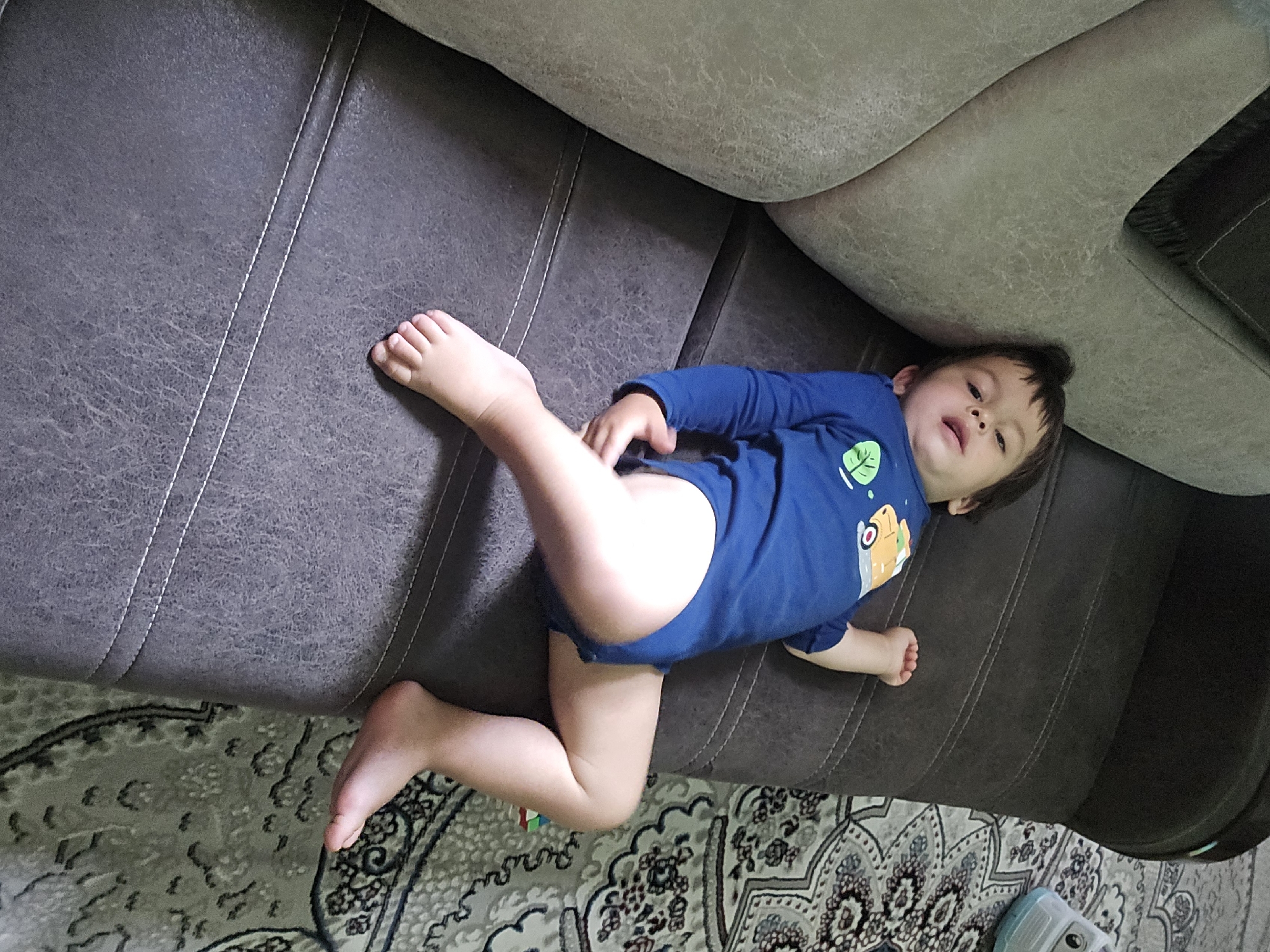 A young child, likely a toddler, lies on their back on a dark gray sofa. The child is wearing a blue long-sleeved bodysuit with a green leaf design on the left chest and a cartoonish yellow vehicle on the right. The child's legs are bent, and their feet are visible. The sofa has white stitching detailing its seams. In the background, a patterned rug with intricate designs in light gray and beige is visible on the floor. The lighting suggests it is daytime, possibly indoors. The child appears relaxed, with their eyes open and mouth slightly ajar, looking upwards. The scene is set in a domestic environment, possibly a living room, in Yakkabog, Uzbekistan. No visible text is present in the image.