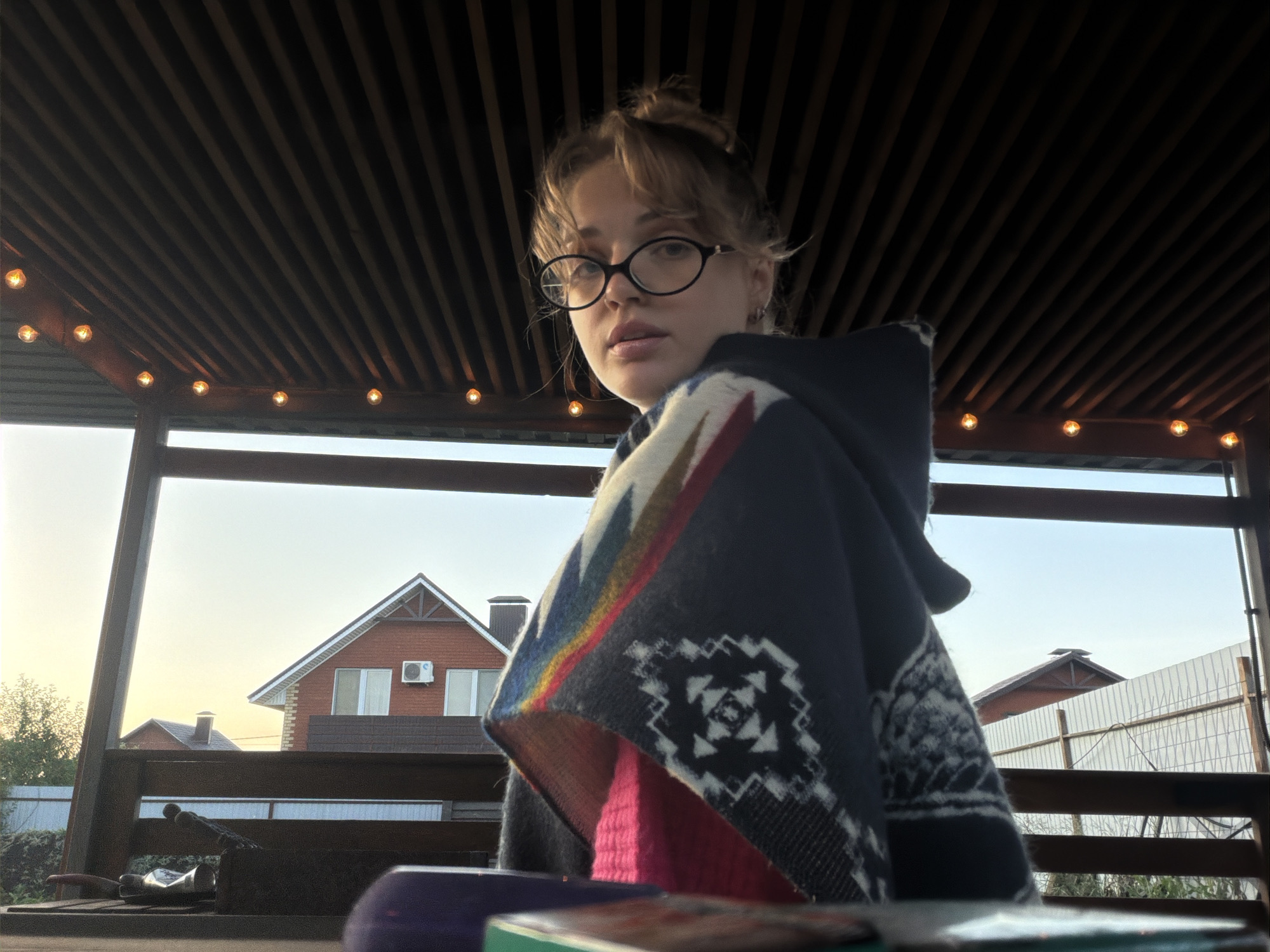 A young woman with light-colored hair tied in a bun and bangs framing her face looks directly at the camera. She wears round, dark-framed glasses and a dark poncho or shawl adorned with colorful geometric patterns in red, blue, white, black, green, and yellow. A bright pink garment is visible underneath the poncho.
She is seated under a dark wooden slatted ceiling, likely part of a porch or gazebo, which features a string of warm, glowing decorative lights. The lighting suggests late afternoon or early evening, with a bright sky visible through the open structure. In the background, a red brick house with white-framed windows and an air conditioning unit is visible, along with another smaller house, trees, and a light-colored fence. Blurry objects, possibly art supplies like a tube of paint and brushes, are visible in the immediate foreground on a surface. The scene portrays a relaxed outdoor setting at dusk in Большая Шильна, Russia. No visible text is present.