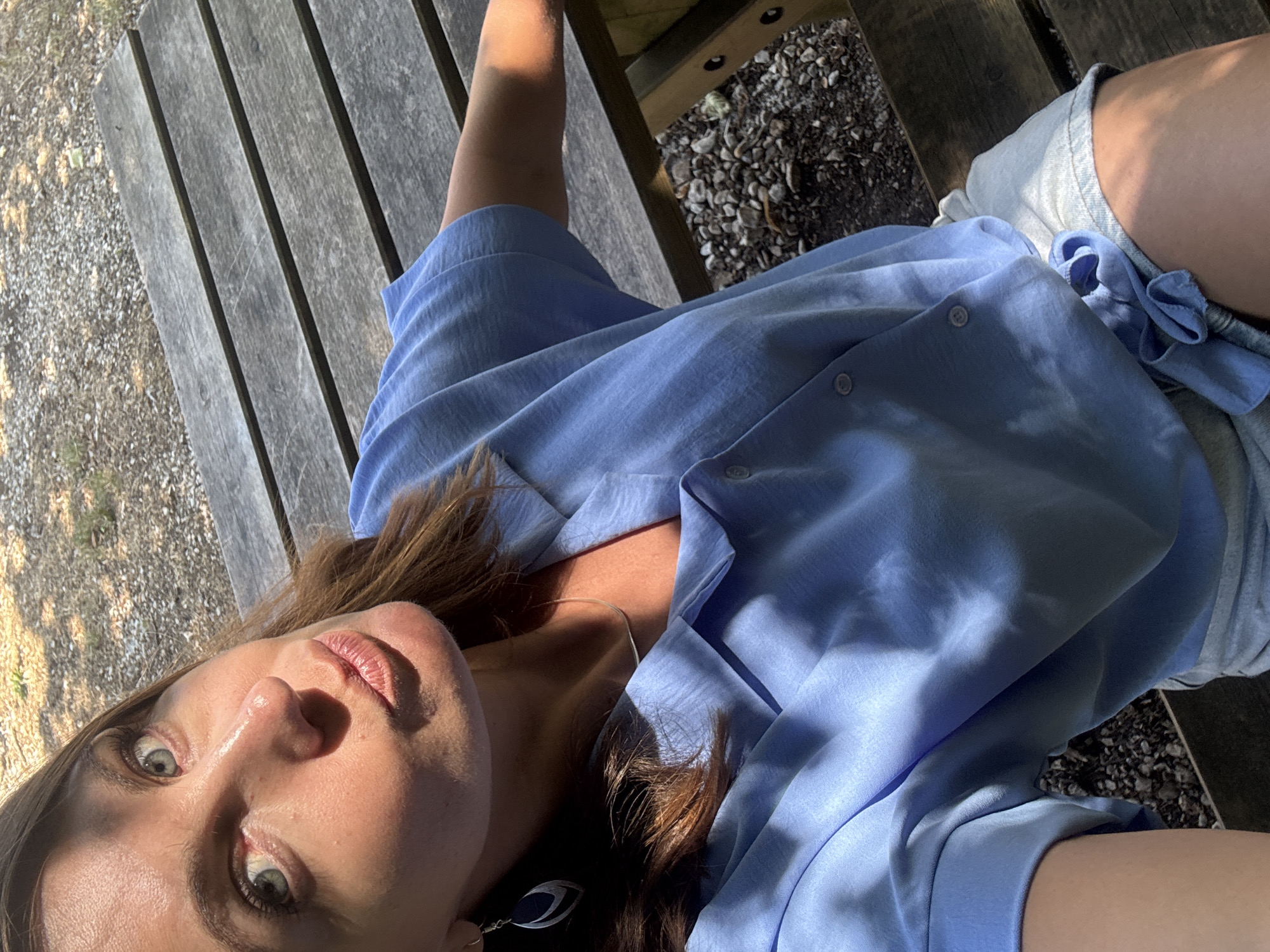 A woman, with light blue eyes and brown hair, is seated on a wooden picnic table outdoors. She is wearing a light blue collared shirt and light denim shorts. Her left arm is resting on the table, and she is looking towards the camera, with her lips pursed. The background shows the texture of the wooden picnic table and gravel. The image appears to be taken during the day, with sunlight casting shadows on the woman and the table. The location is not specified in the image nor its metadata, and therefore cannot be identified as Coteaux du Lizon, France.