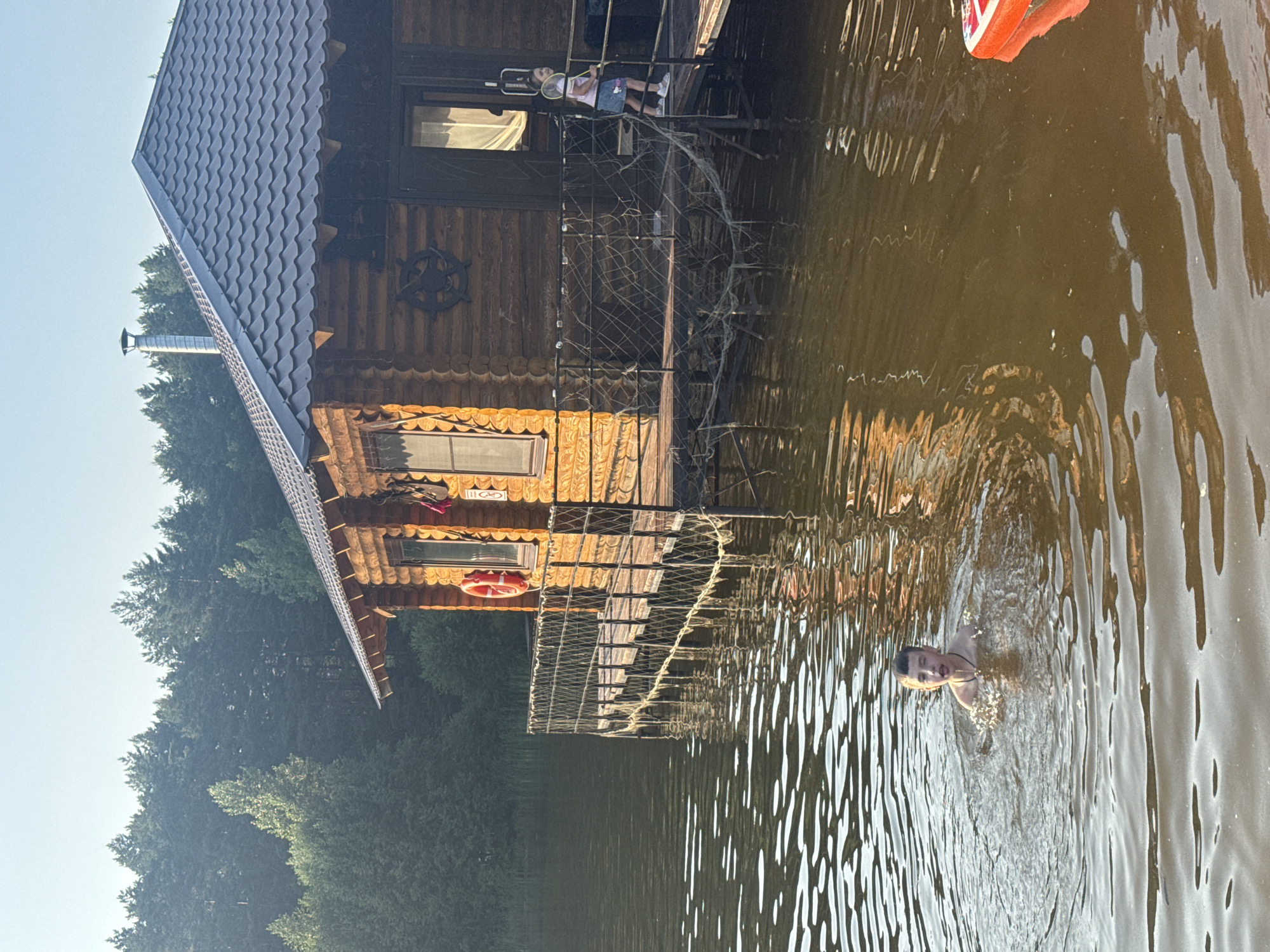 A boy is swimming in a lake in front of a wooden house. The house is on stilts and has a deck with a railing. There is a girl on the deck of the house holding a badminton racket. The lake is surrounded by trees. The sun is setting, casting a warm glow on the scene.