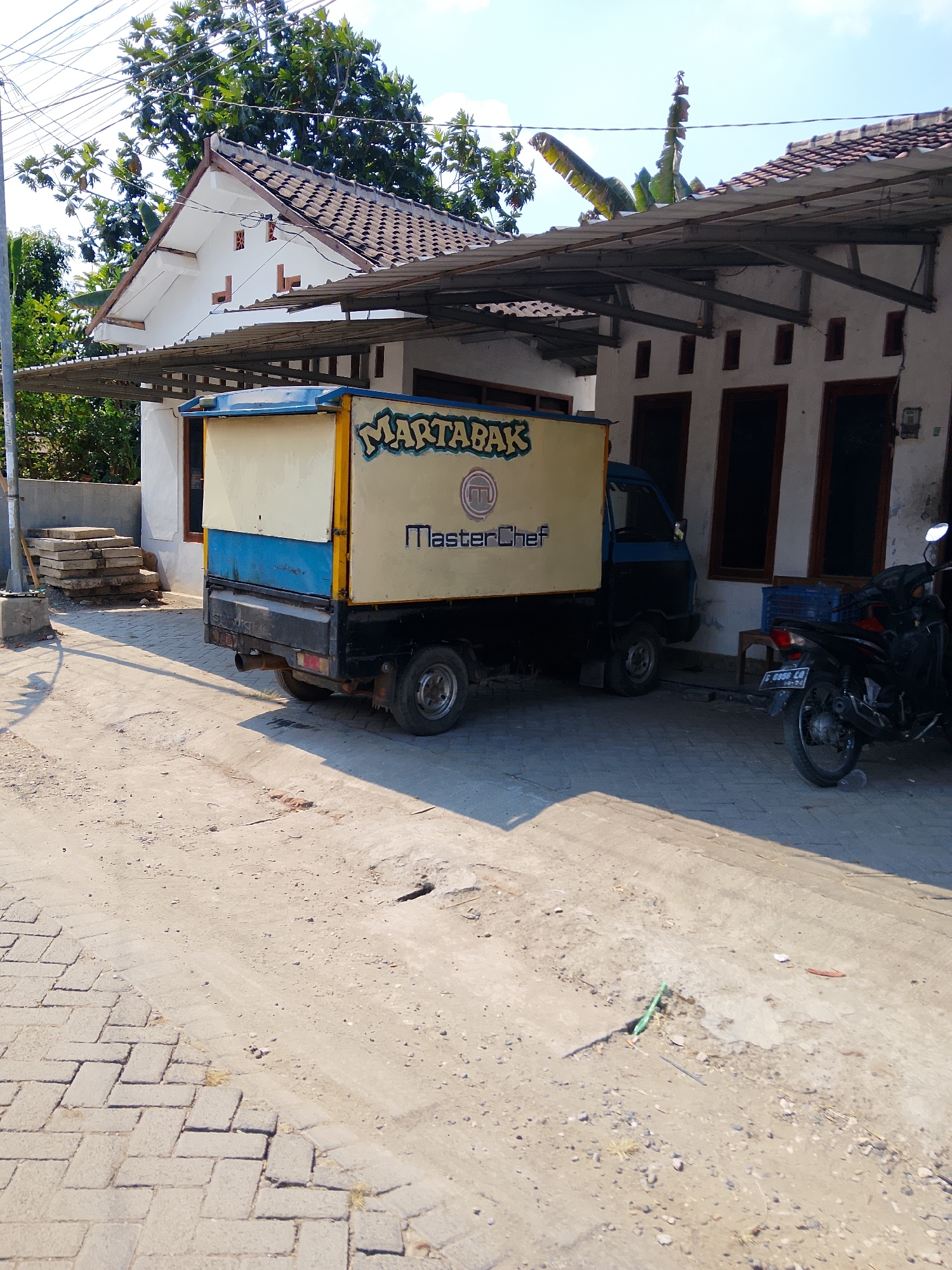 This image depicts a street scene in Padangan, Indonesia. In the foreground, a cobblestone pathway borders a dusty dirt road. A light blue and yellow truck, branded with "MARTABAK MASTERCHEF," is parked on the dirt road. The truck is positioned in front of a white building with a covered awning extending from its roof. To the right of the truck, a dark-colored motorcycle is parked. The background features lush green trees and a clear blue sky.