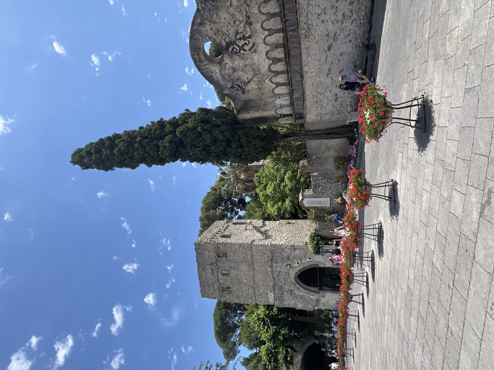 This media file captures a sunny day in Ravello, Italy, showcasing a charming architectural scene. The foreground is dominated by a cobblestone plaza, where elegant planters filled with vibrant red and white flowers line the pathway. These planters are supported by ornate black metal stands, casting distinct shadows on the ground, indicating the time of day is likely midday given the bright sunlight.
In the midground, a stone tower with an arched entrance stands prominently. The tower exhibits weathered stone construction, hinting at its historical significance. Beyond the tower, lush green trees and foliage create a natural backdrop. To the left, a tall, slender evergreen tree reaches towards a bright blue sky dotted with fluffy white clouds.
Further into the scene, an ancient stone structure with decorative elements and a circular opening is visible, suggesting a historical building or ruin. Two individuals are seated on a stone ledge in front of this structure, appearing relaxed and enjoying the view. Scattered throughout the plaza, other visitors can be seen, some walking, others pausing to admire the surroundings, contributing to a peaceful, leisurely atmosphere. The overall impression is one of a beautiful and historic tourist destination bathed in warm sunlight. No visible text is discernible in the image.