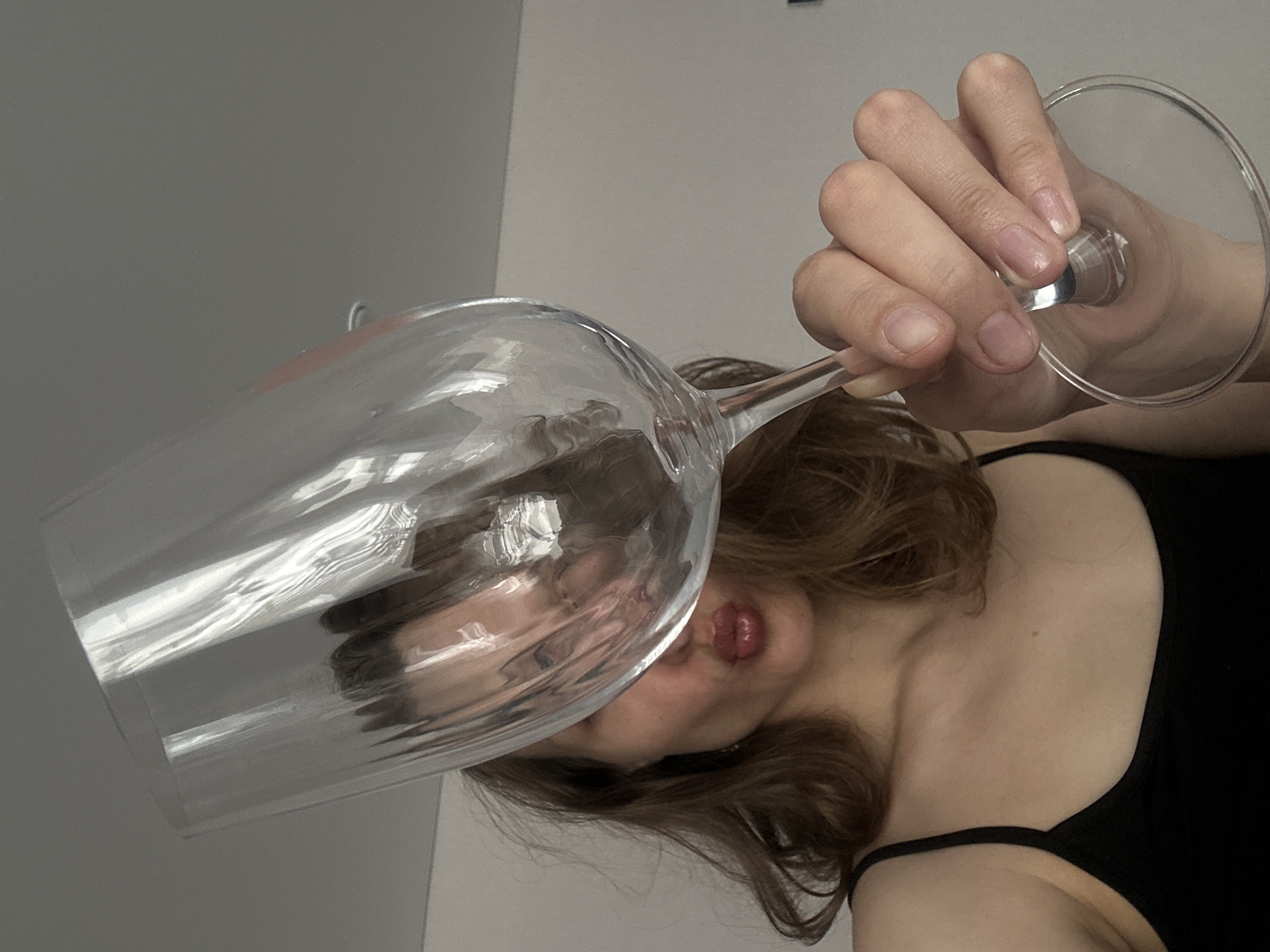 A woman is holding a wine glass up to her face. The woman has brown hair and is wearing a black top. The background is a plain gray wall. The location is unknown and is not evident from the image. The glass is empty. Her face is distorted through the glass. The scene is indoors.