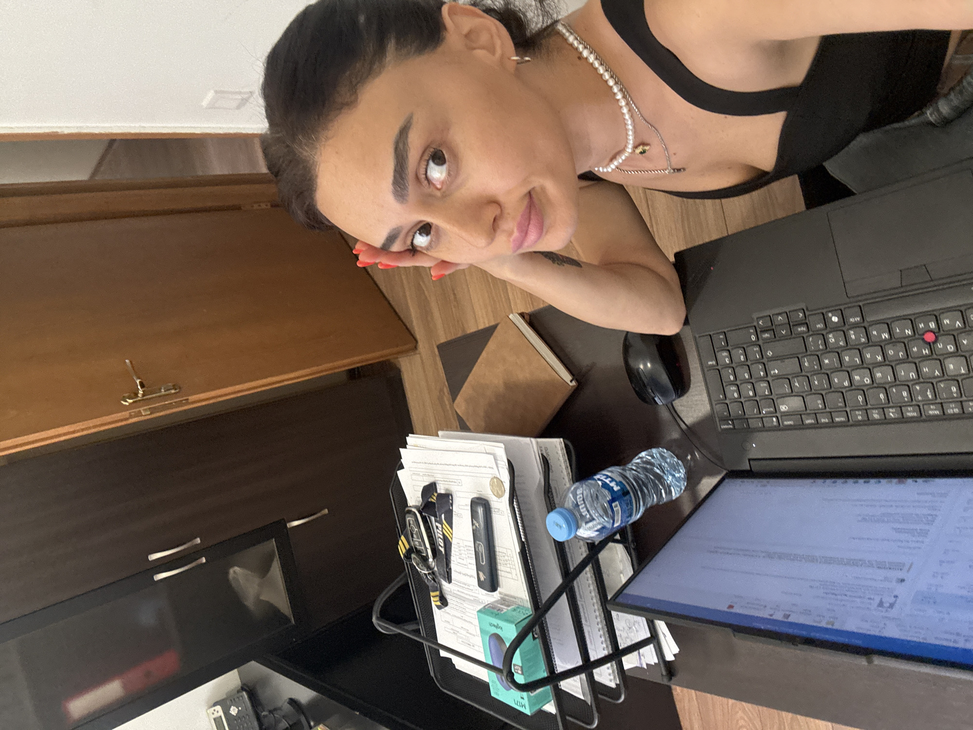 This is a vertical, eye-level, medium shot of a woman sitting at a desk in what appears to be an office or home workspace. The woman is in the foreground, leaning her head on her hand and looking up towards the camera with a slight smile. She has dark hair pulled back and is wearing a black top and pearl necklaces.
On the desk, a black laptop is open with its screen visible, showing text. Next to the laptop is a computer mouse and a clear plastic water bottle with a blue cap. In front of the laptop, there is a metal document holder filled with papers, a pen holder with some pens, and a small green box. A notebook lies to the side of the document holder.
In the background, a wooden cabinet or door is visible, suggesting an indoor setting. The lighting is bright, and the overall mood is casual and perhaps a bit playful, given the woman's expression. The time of day is not explicitly indicated but the bright lighting suggests it could be daytime. Visible text includes "PIL" on a lanyard, and some text on the water bottle and the green box, as well as text on the laptop screen and documents. The location context is Tbilisi, Georgia.