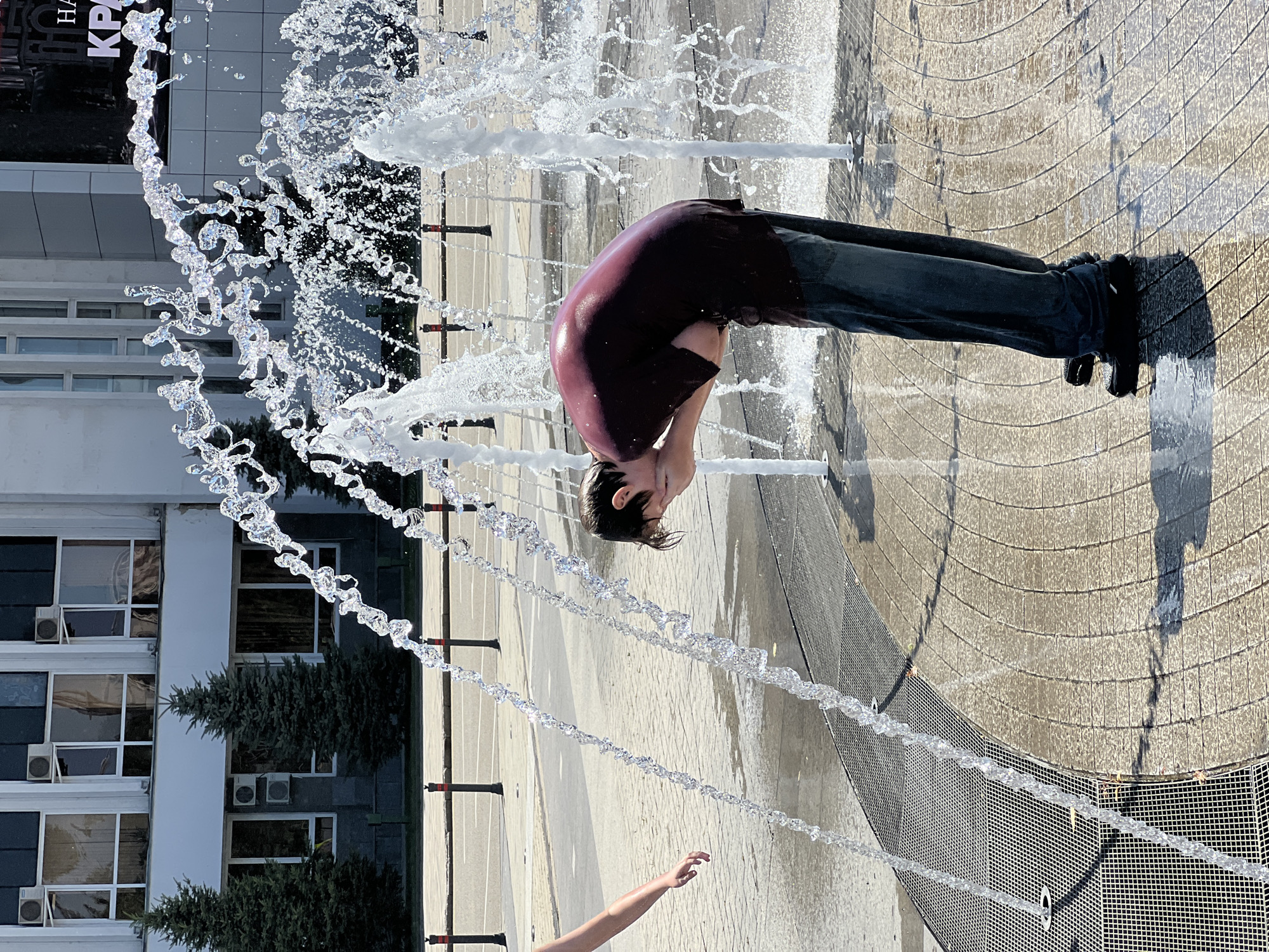 A young boy is playing in a public fountain. He is wearing a dark maroon t-shirt and dark blue jeans. His head is bent down, and he is splashing water on his face with his hands. The fountain is located in a paved area with a textured surface, likely a city square or park. The background shows a building with large windows and some trees. The image is taken from a low angle, capturing the dynamic spray of the water.